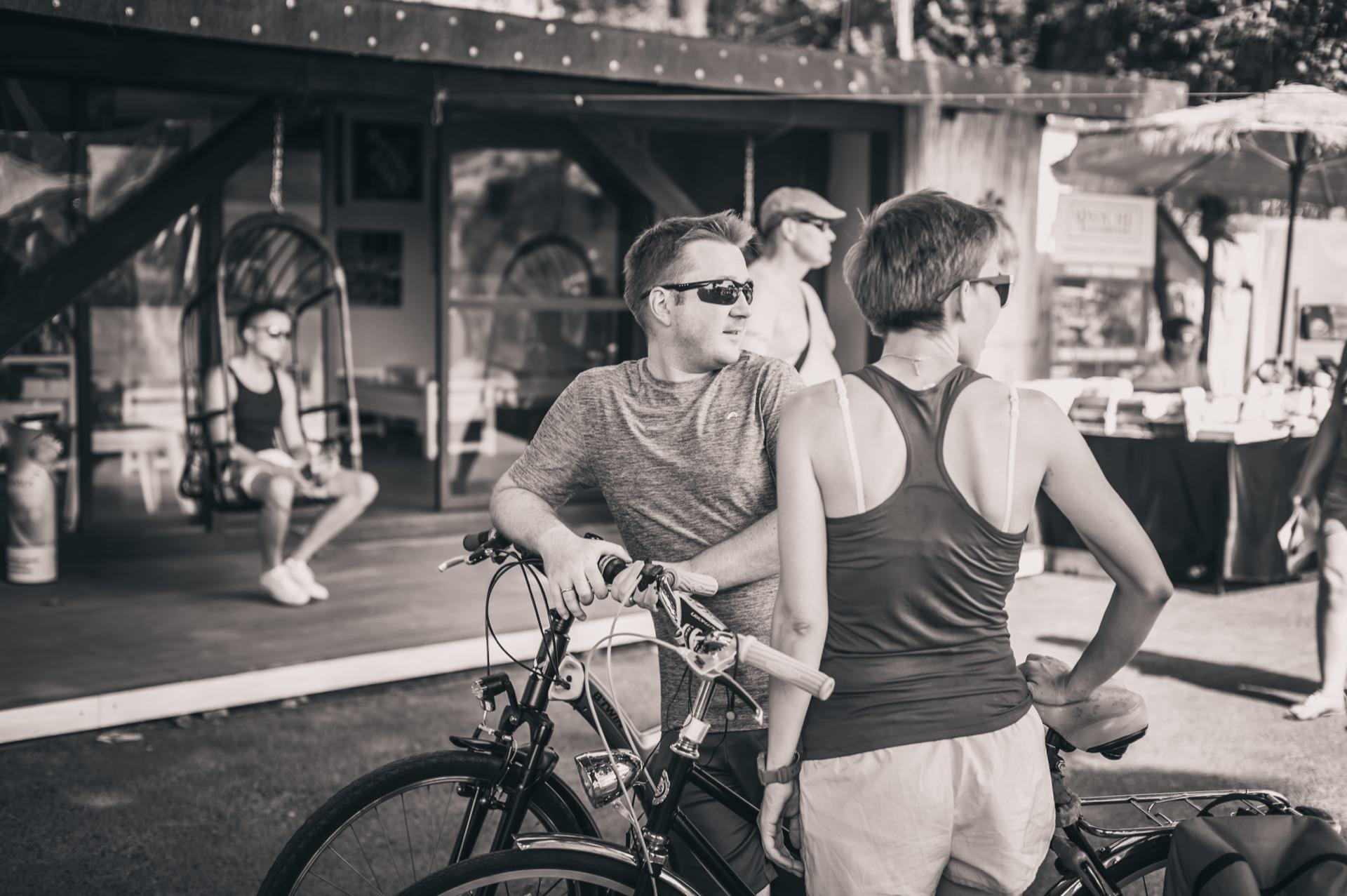 A black-and-white photo of two people on bicycles, wearing everyday clothes and sunglasses, standing in front of a wooden structure in a bustling market. One person is sitting on a swing. The scenery appears to be a bustling place, capturing the essence of the photo fair.  