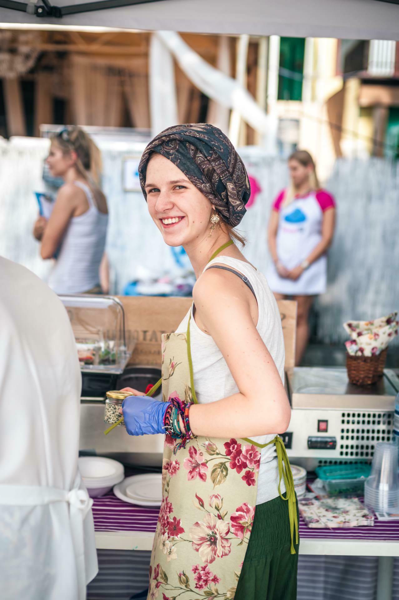 A woman wearing a floral apron and a headscarf smiles as she works at an outdoor food stand. She prepares food with gloves on, standing by kitchen equipment and a table laden with dishes. In this photo essay from the fair, other people are blurred in the background, indicating a busy scene.  