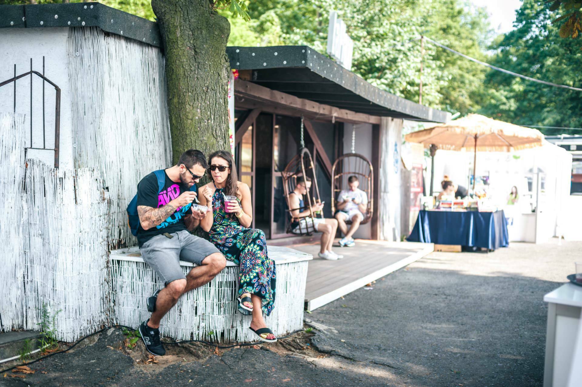 A man and a woman sit on a white wooden shelf under a tree, engrossed in phones or drinks. The woman is dressed in a floral dress and sunglasses, while the man is in casual attire. In the background, several people are relaxing on this sunny day near a small hut and a shady spot - capturing these moments as part of our photo report of the fair.  