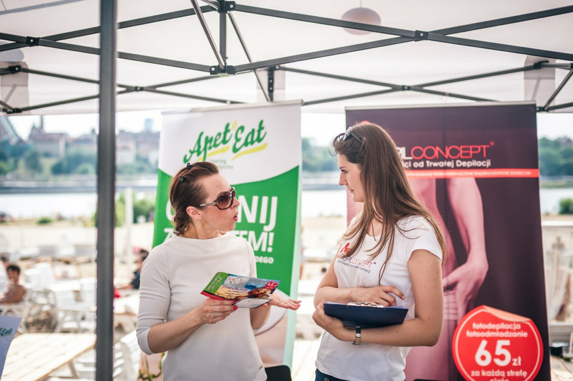 Two women are standing and talking under a canopy at an outdoor event. One is holding a brochure and the other has a clipboard. Behind them are two large promotional banners with text and images - part of the Photo Fair. In the background you can see tables, chairs and the riverbank.   