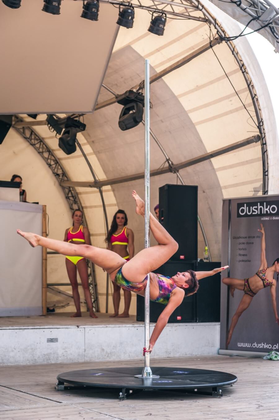 Kobieta wykonuje na scenie powietrzny taniec na rurze, trzymając rurę jedną ręką i jedną nogą wyciągniętą na zewnątrz. Nosi kolorowy strój sportowy. W tle stoją dwie kobiety w podobnych strojach, z widocznym na scenie banerem i zestawem DJ-a, rejestrując tętniącą życiem fotorelację z targów.