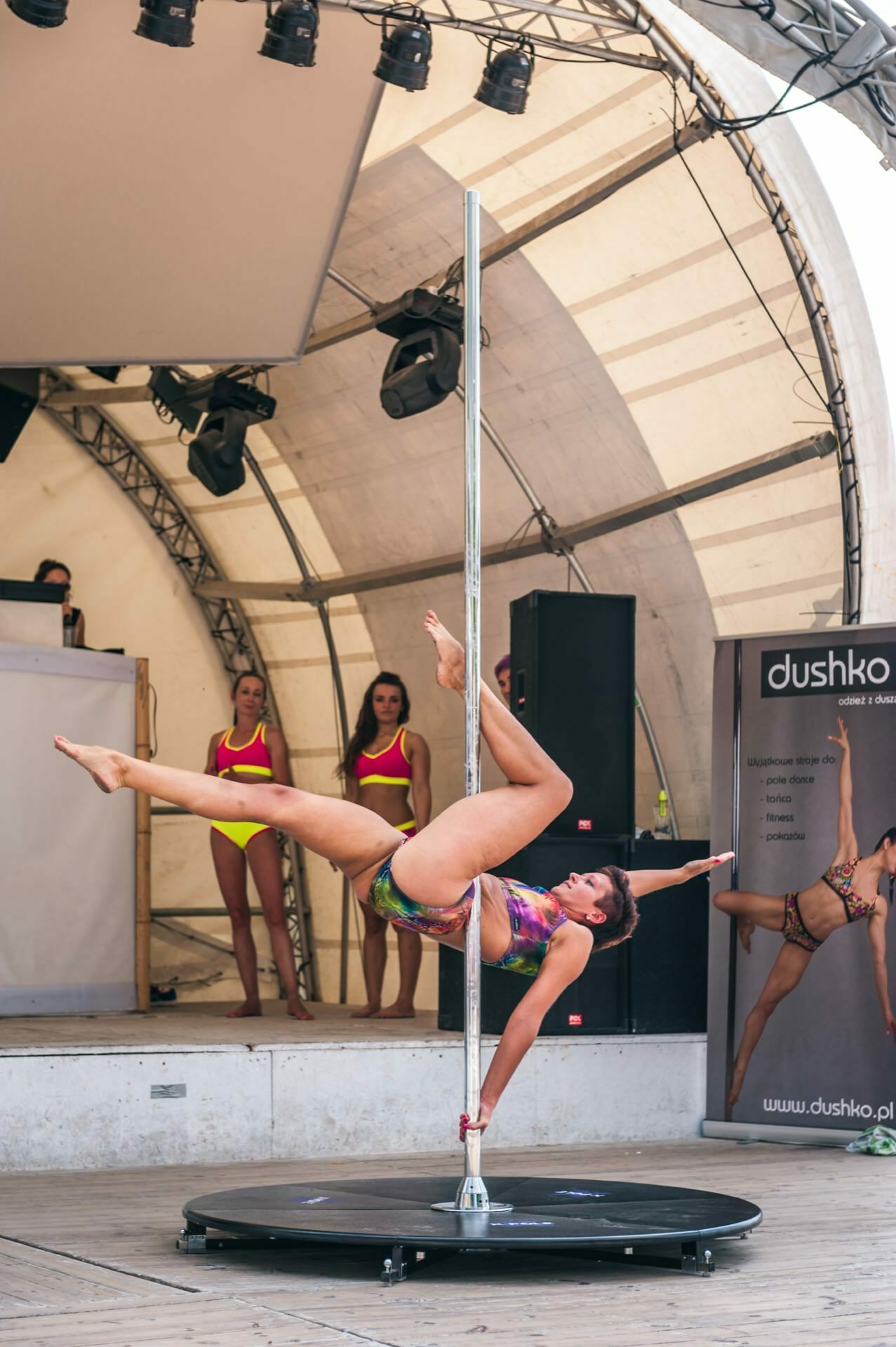 A woman performs an aerial pole dance on stage, holding the pole with one hand and one leg extended outward. She wears a colorful sports outfit. Two women in similar outfits stand in the background, with a banner and DJ set visible on stage, capturing a vibrant photo-op of the fair.  