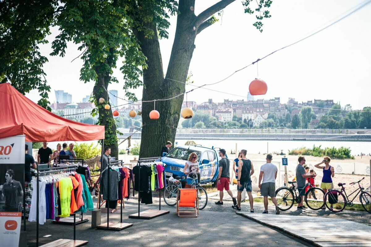 Rynek na świeżym powietrzu pod dużymi drzewami obok rzeki. Na pierwszym planie namiot z kolorowym strojem sportowym na stojakach. Ludzie przeglądają rynek i jeżdżą na rowerach, tworząc tętniącą życiem fotorelację z targów. W tle widać panoramę miasta z zawieszonymi nad głową latarniami.