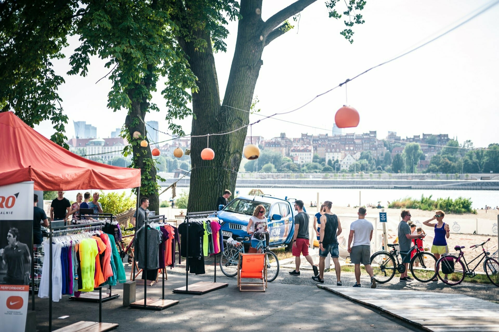 Outdoor market under large trees next to the river. In the foreground is a tent with colorful sportswear on racks. People browse the market and ride bicycles, creating a vibrant photo-op of the fair. In the background you can see the city skyline with lanterns hanging overhead.   