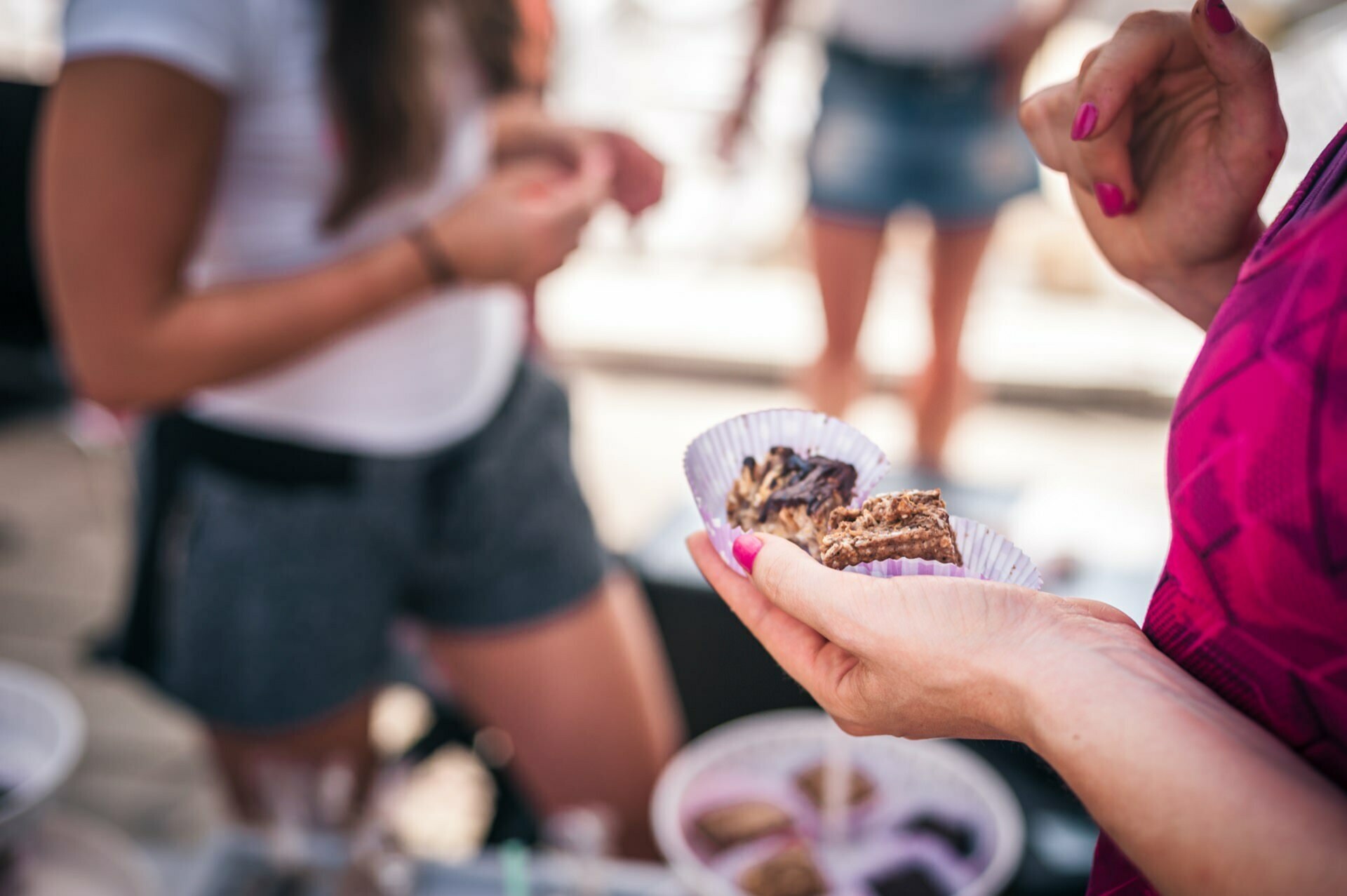 A person in a pink shirt is holding a paper cup with pieces of baked goods, while another person in the background is wearing a white top and gray shorts. A table set with a variety of baked goods can be partially seen - perfectly captured in the photo report of the fair from the outdoor event. 