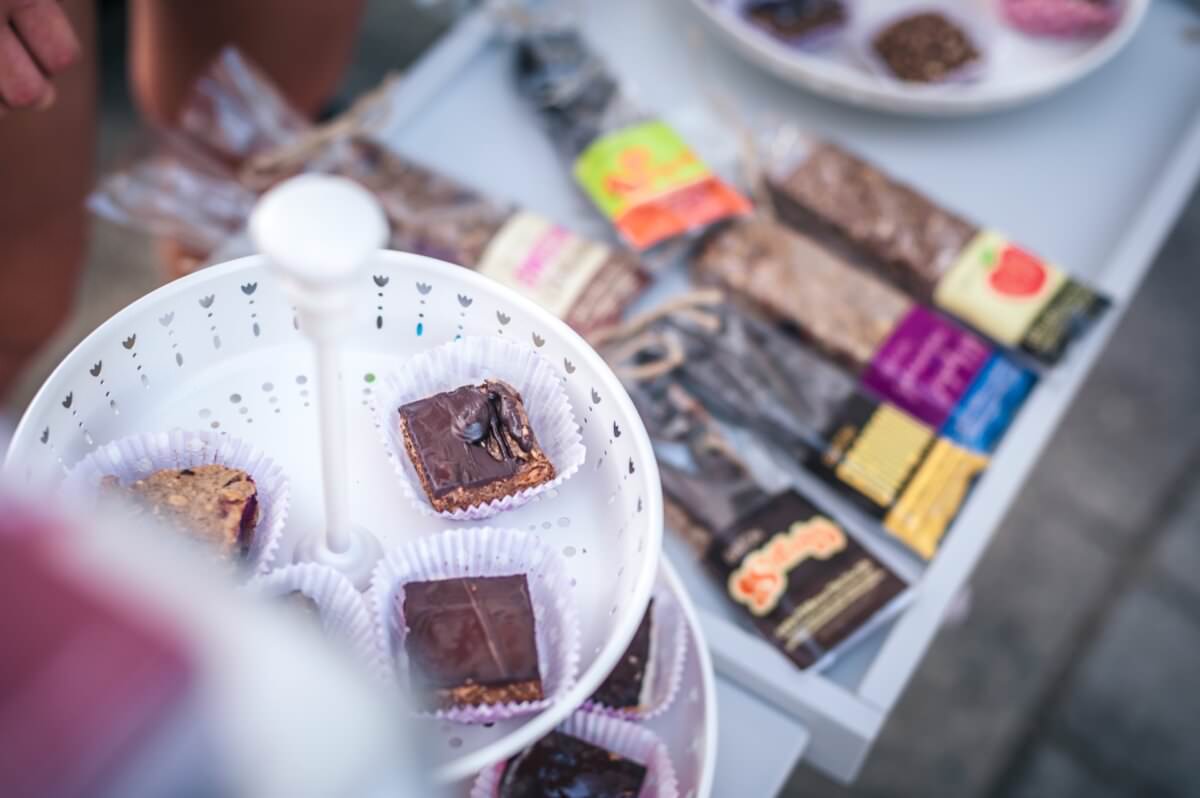 Wyświetlany jest trójpoziomowy biały stojak z różnymi smakołykami w czekoladzie, niektóre z wyjętymi kęsami. W tle na białym stole leży asortyment owiniętych batonów energetycznych. Scena wydaje się pochodzić z imprezy plenerowej, uwieczniając żywą fotorelację z targów.