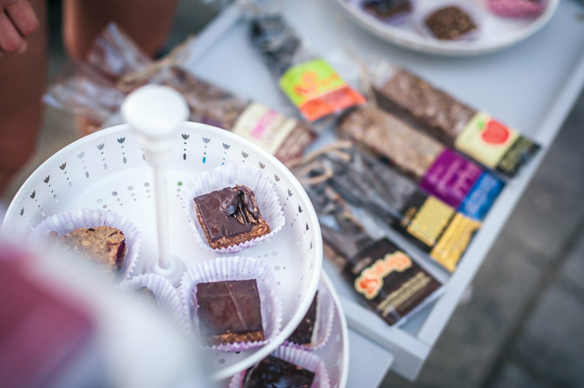 Displayed is a three-tiered white stand with a variety of chocolate-covered treats, some with bites taken out. In the background, an assortment of wrapped energy bars lie on a white table. The scene appears to be from an outdoor event, capturing a lively photo-op of the fair.  