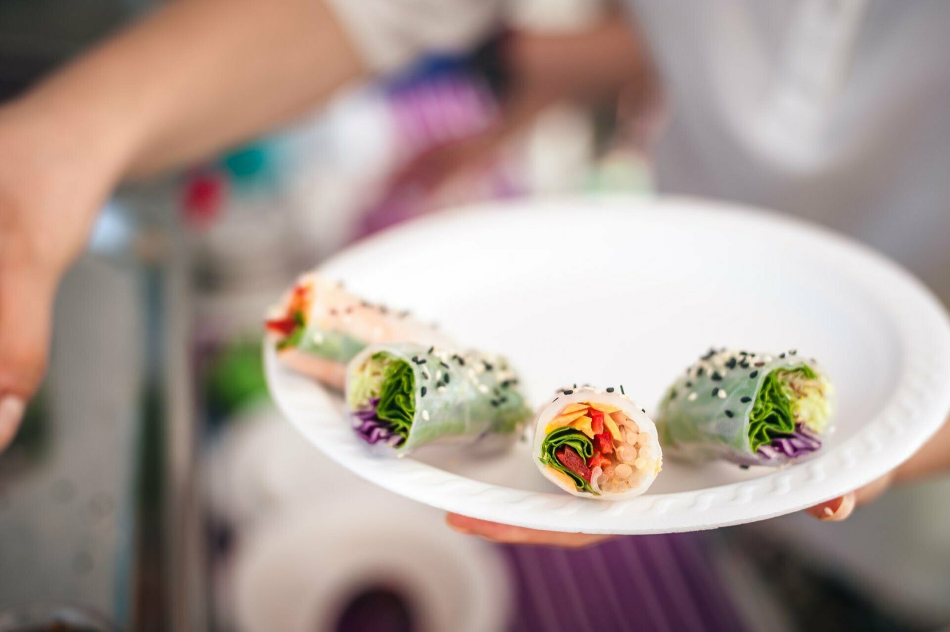 A white plate with four fresh spring rolls cut in half, revealing vibrant fillings, including sliced vegetables and veggies. The spring rolls are sprinkled with black sesame seeds, and in the background you can see a hand reaching toward the plate - the perfect shot for a *fair photo story. 