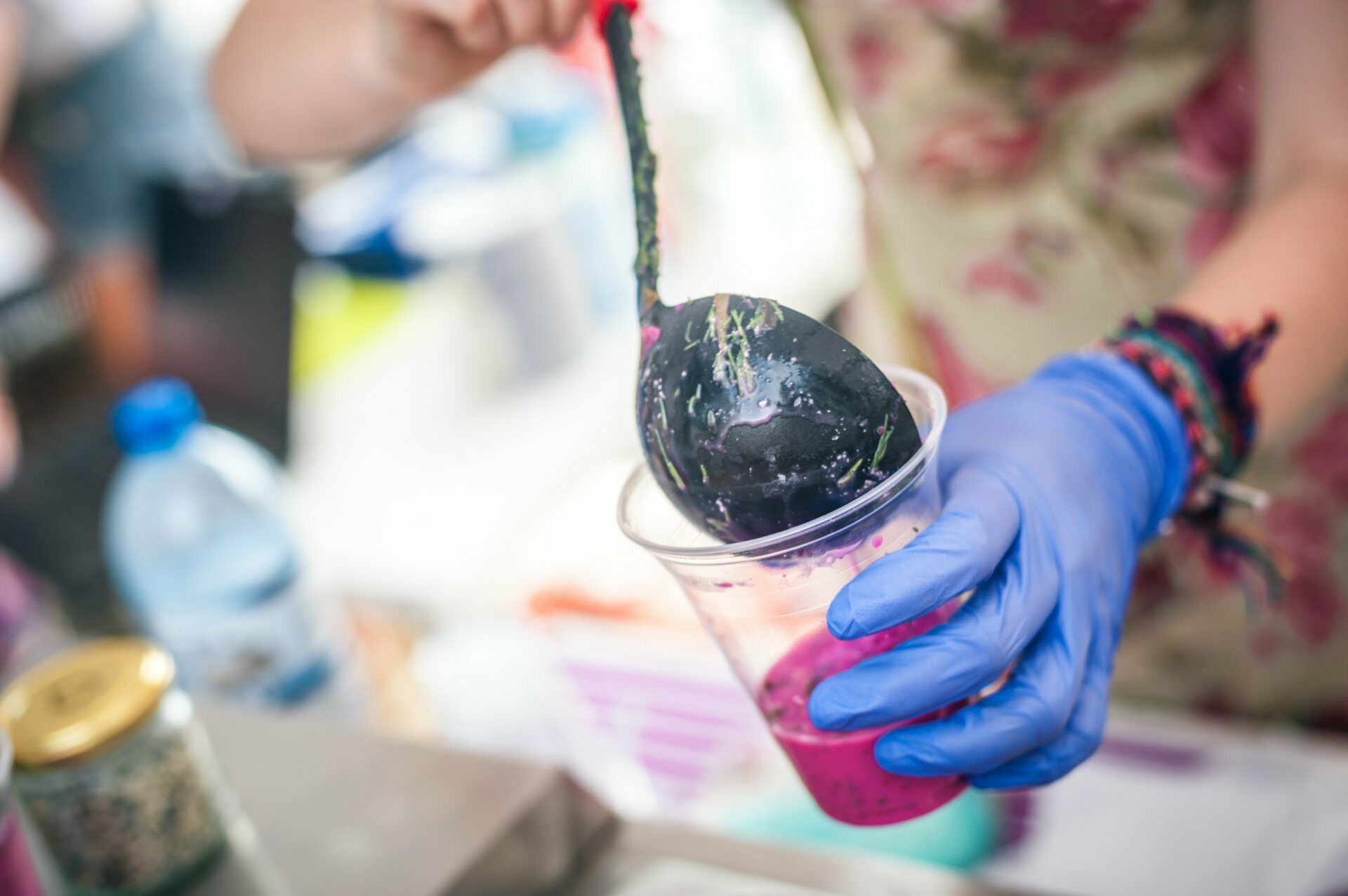 A photo essay from the fair: A person wearing blue gloves pours a purple liquid from a ladle into a clear plastic cup. The background is blurred, but a bottle of water and a small jar are visible. A person wearing an apron with a floral pattern adds to the colorful scene.  