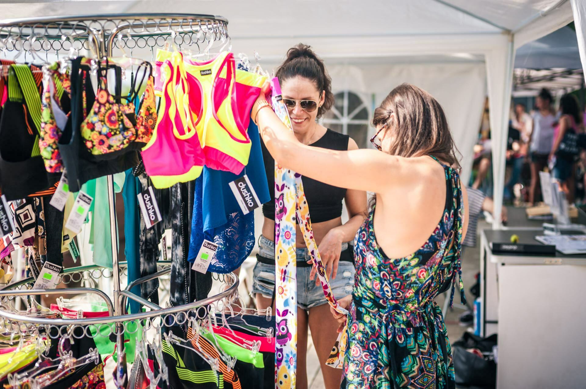 Two women are looking at colorful sports bras and leggings on a round rack at an outdoor market. The woman on the left is wearing sunglasses and a black top, while the woman on the right, also wearing sunglasses, is wearing a patterned dress. A white canopy tent is visible in the background, capturing the moment of the photo fair.  