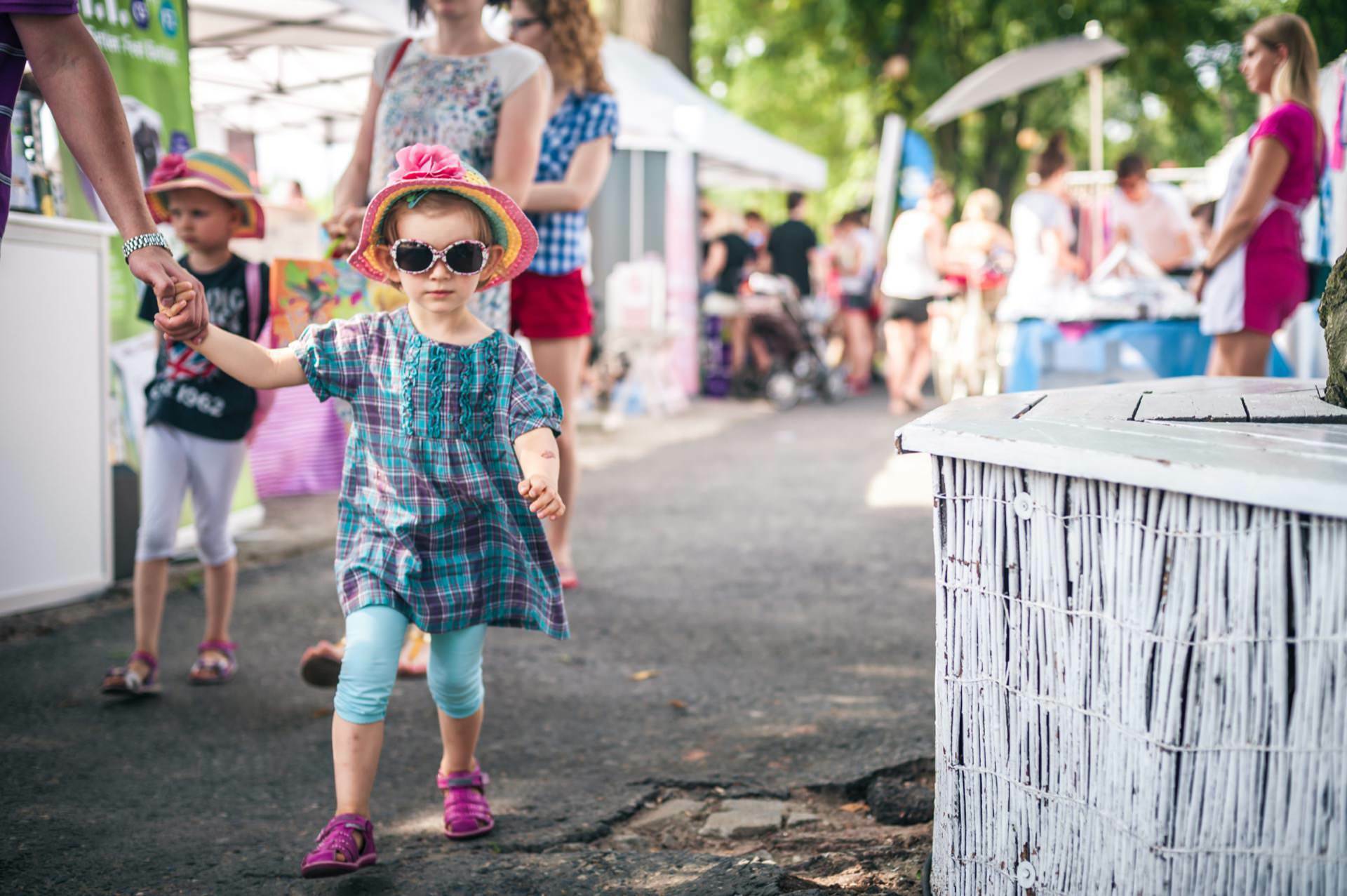 A young child wearing sunglasses, a colorful hat and a checkered dress walks arm-in-arm with an adult at an outdoor event. He is followed by another child. Blurry people and stalls can be seen in the background, creating a vivid photo-reportage of the fair.  