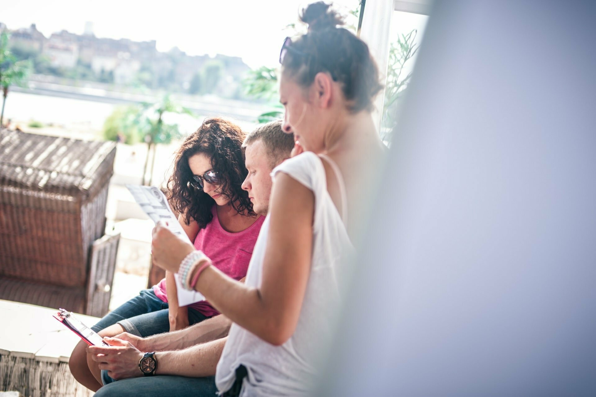 A group of three people are sitting together enjoying a sunny day. One person with curly hair and sunglasses is smiling, another is holding a piece of paper and the third is looking at a tablet. They are sitting outdoors with a blurry background - perfect for a photo fair.  