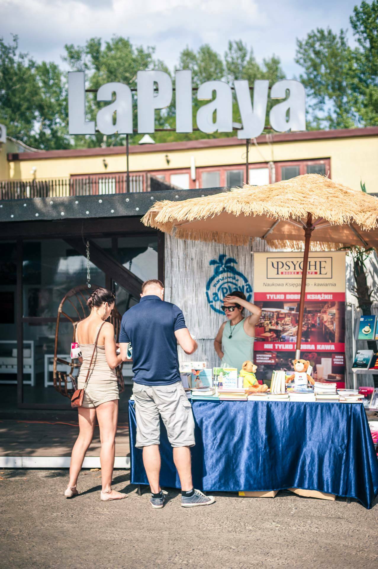 A small outdoor booth at the event under a sign reading "LaPlaya" hosts three people, including a woman in a hat behind a table, handling the items on display. A colorful booth covered with a blue tablecloth and sheltered by a straw umbrella creates a picturesque photo-op of the fair. 