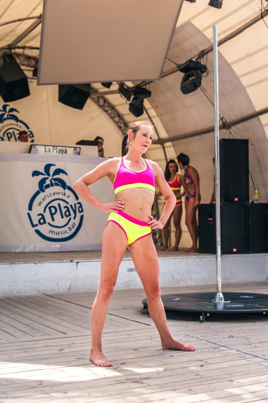 Kobieta ubrana w różowo-żółty dwuczęściowy strój sportowy stoi pewnie na drewnianej scenie podczas imprezy plenerowej pod dużym namiotem. Inne kobiety w podobnych strojach znajdują się w tle w pobliżu szyldu „La Playa” i stoiska DJ-a, rejestrując tętniącą życiem scenę fotorelacji z targów.