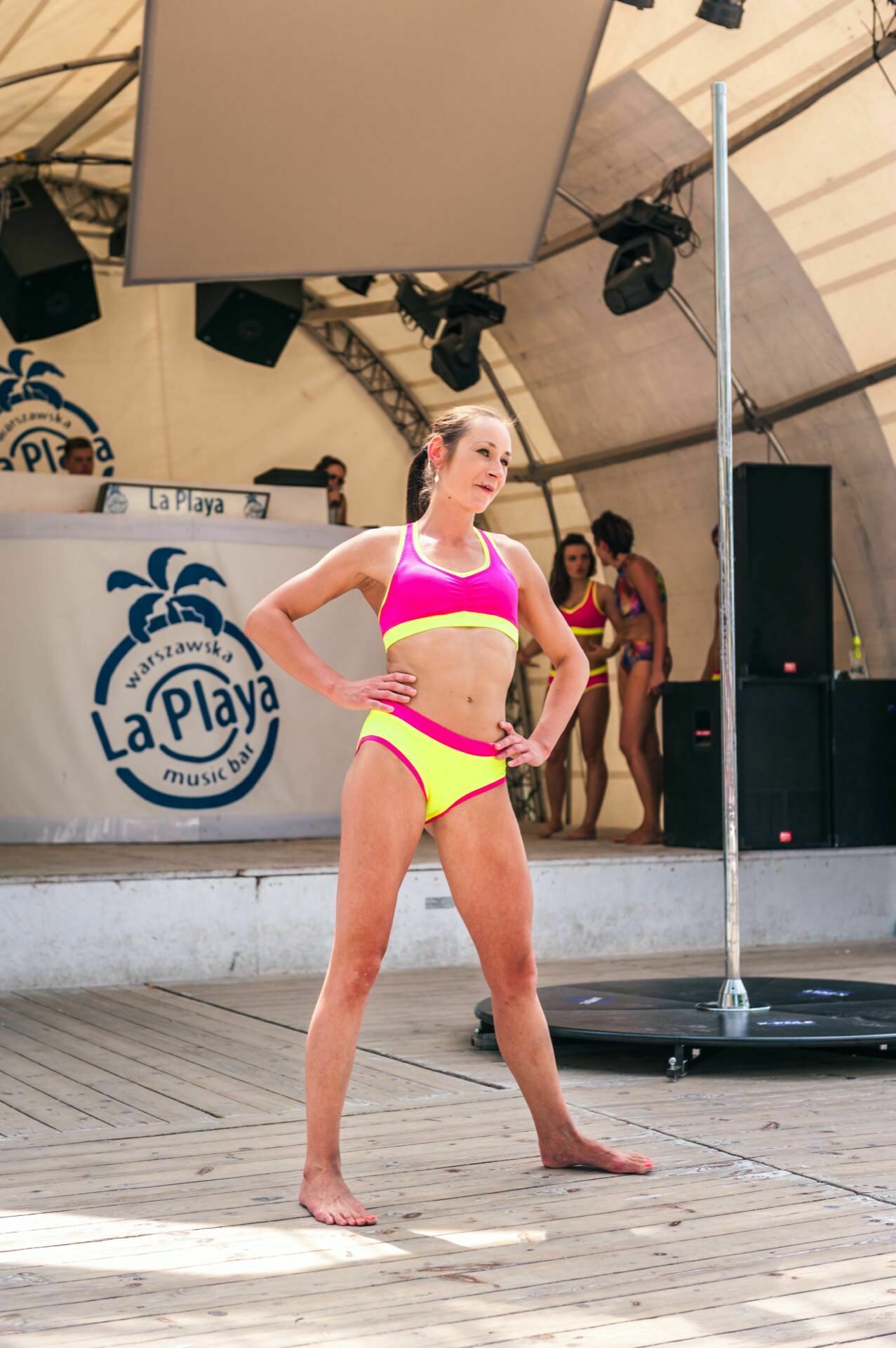 A woman dressed in a pink and yellow two-piece sports outfit stands confidently on a wooden stage during an outdoor event under a large tent. Other women in similar outfits are in the background near the "La Playa" sign and DJ booth, capturing the bustling scene of the photo fair. 