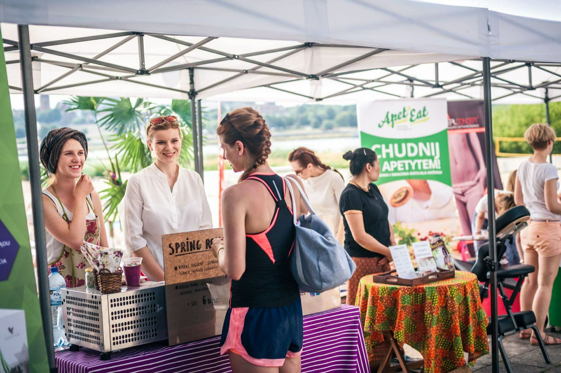 Photo report from the fair: People at a stall under tents on a sunny day. A woman in a pink T-shirt and shorts chats with two smiling vendors behind a table covered with a purple striped tablecloth. Various items are presented on the table with signage in the background.  