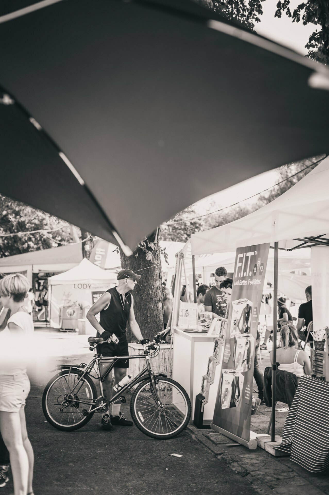 Black and white photo of an outdoor market. A man stands with his bicycle and looks at a stand with the sign "F.I.T." nearby. In the background you can see other booths and people, with large umbrellas providing shade - a truly captivating photo of the fair.  