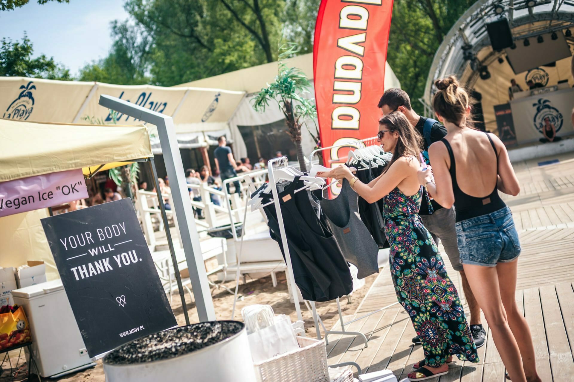 People browse through clothes at a market or outdoor festival on a sunny day. One woman is wearing a floral dress, another a black T-shirt and denim shorts. A large red "DUDADO" flag placed next to the inscription "Your body will thank you" catches the eye. In the background, there are trees and tents - perfect for the Photo Fair.   