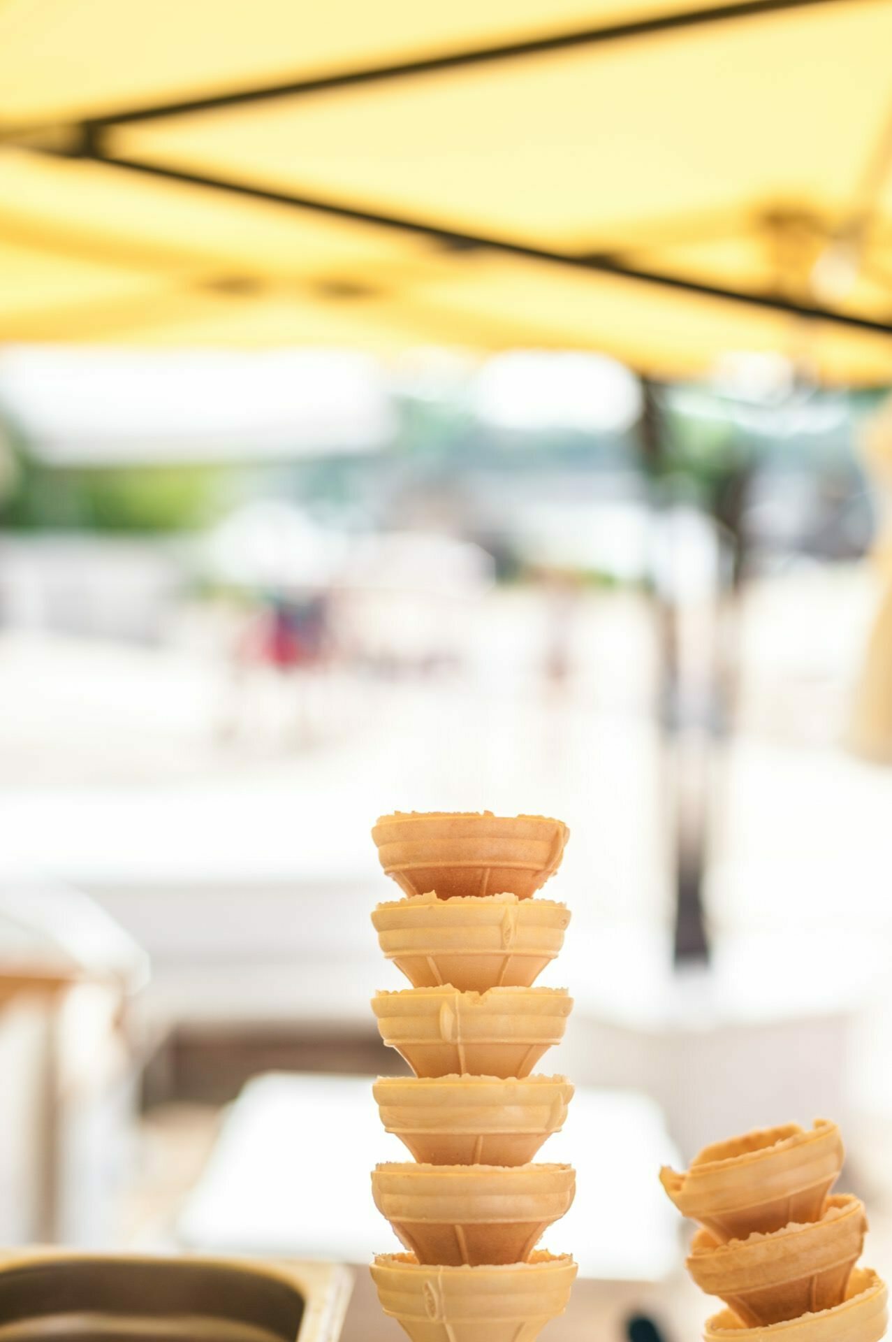 A stack of empty waffle ice cream cones is displayed under a bright yellow canopy in an outdoor setting. The background, gently blurred, depicts a sunny setting, probably by the beach, with people walking in the distance. This scene looks as if it could be part of a photo essay of the fair.  