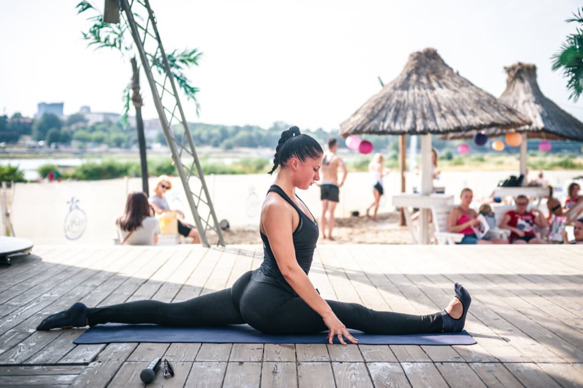 Kobieta w czarnym stroju do ćwiczeń wykonuje szpagaty na macie do jogi na świeżym powietrzu. W tle widać plażę z chatkami i innymi ludźmi odpoczywającymi i wchodzącymi w interakcje, co przypomina fotorelację z targów. W oddali widać palmy i brzeg rzeki.