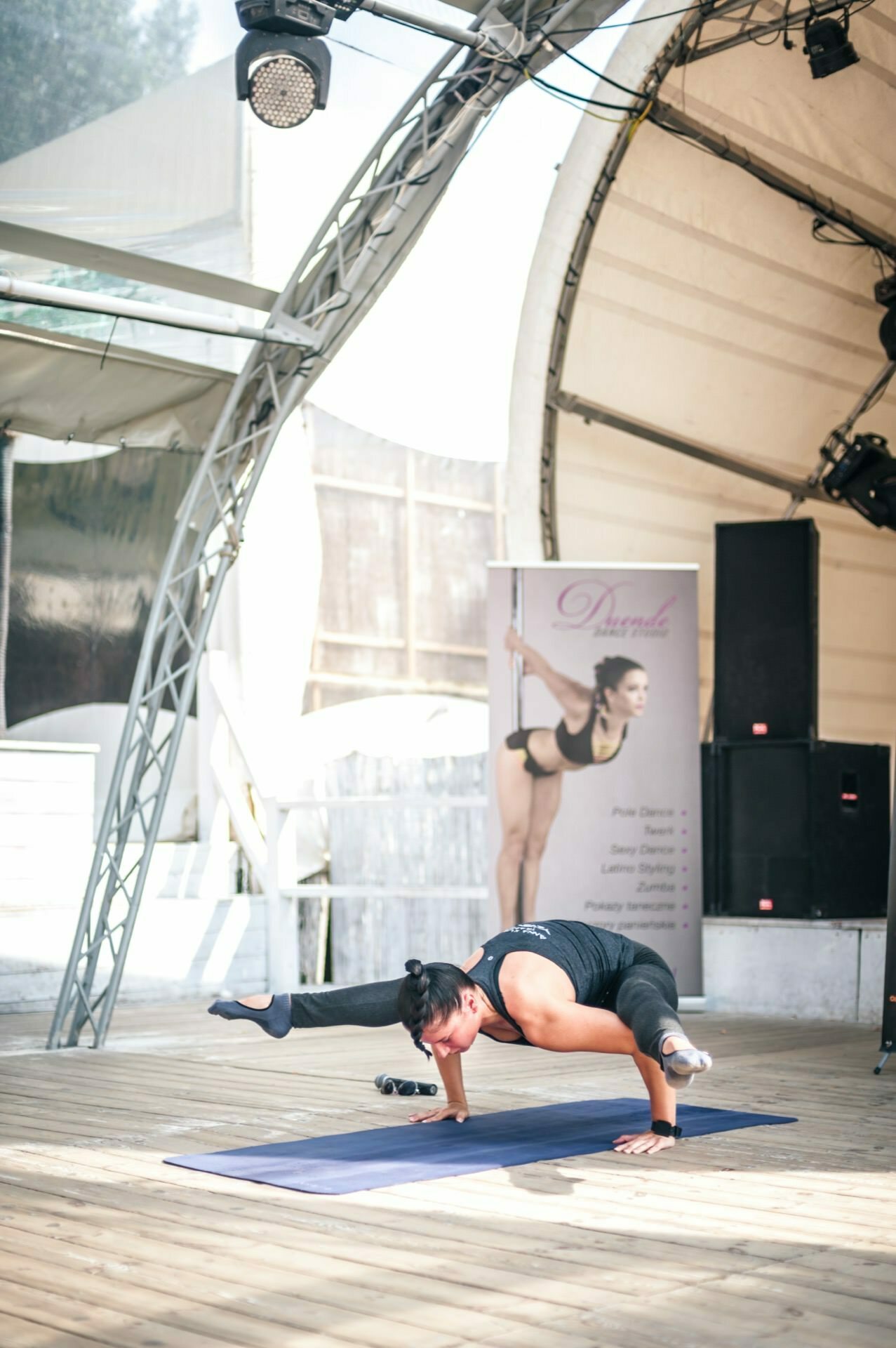 A person dressed in black sportswear performs a challenging yoga pose, balancing on his arms with his legs stretched out to the sides. They are sitting on a blue yoga mat in a covered outdoor space during a photo opportunity. A large poster and sound equipment are visible in the background.  