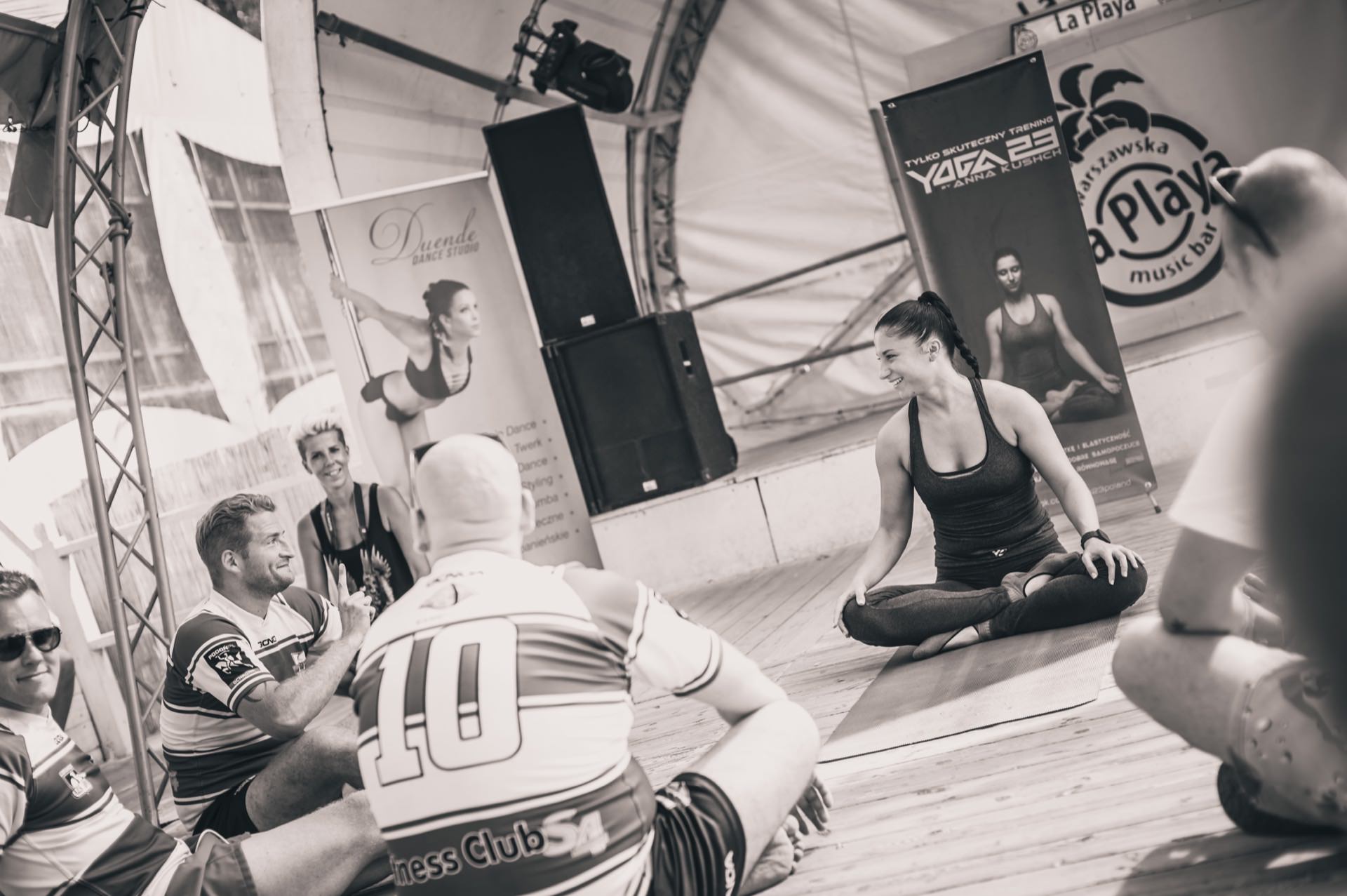 Black and white image of yoga class in session. Several people sit on the floor in a large, bright tent. The instructor sits on a mat with her legs crossed, facing the students, who watch her intently. Banners promoting yoga and fitness surround the space, beautifully capturing the photo-op.   