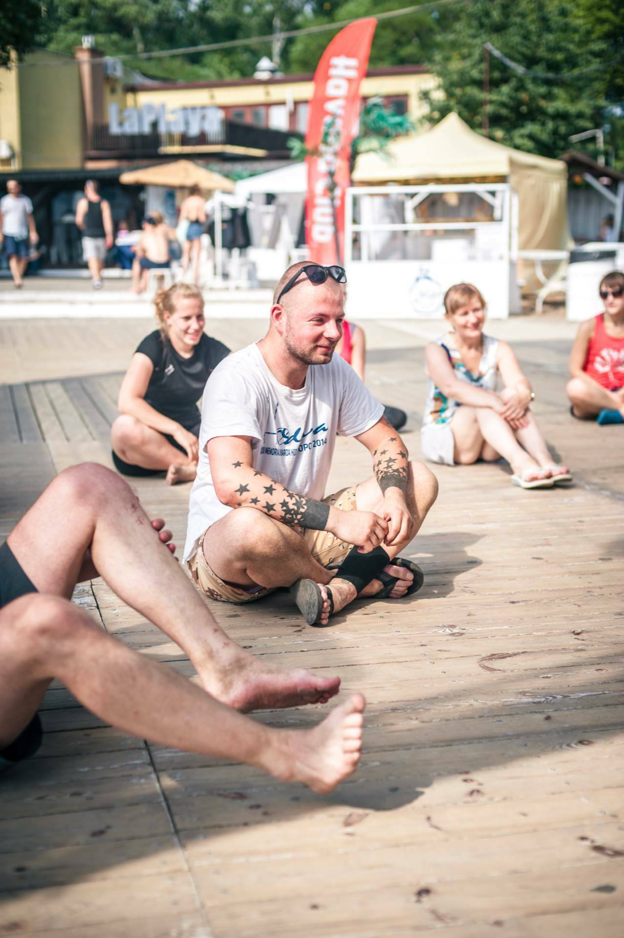 A group of people are sitting on an outdoor wooden terrace. The central figure is a man dressed in a white T-shirt, beige shorts, sandals and sunglasses on his head. The people around him are also sitting casually, some smiling, in a relaxed, sunny environment - perfect for a photo fair.  