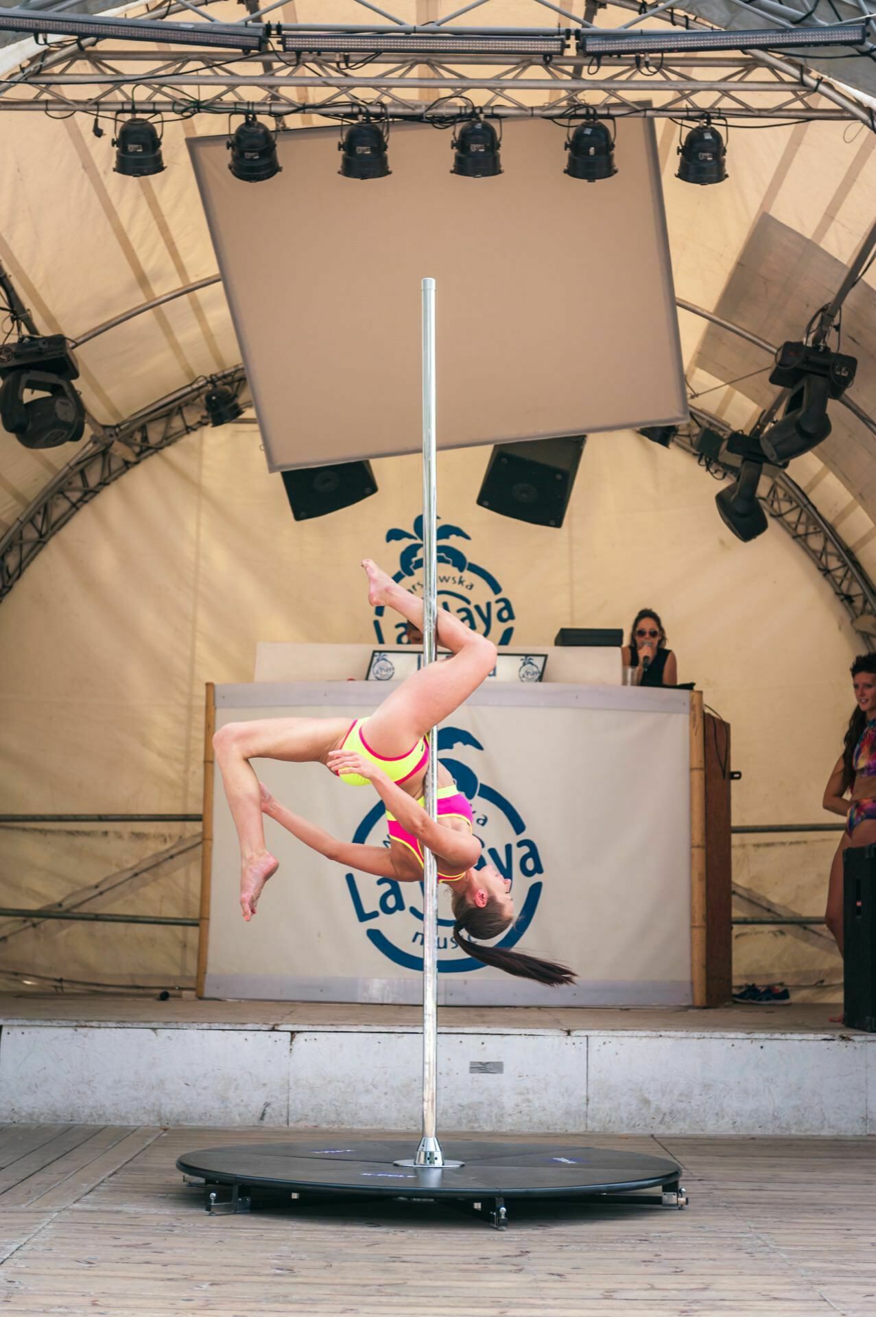 A person in brightly colored sportswear performs an advanced upside-down pole dance on a stage under a tent. In the background you can see a DJ stand with someone behind him and a large sign with the name "La Savaya" and a palm tree logo, capturing the essence of the "Photo Fair". 