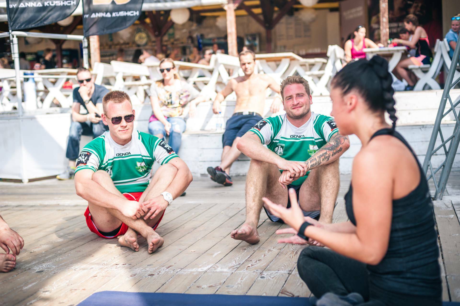 A woman in a black T-shirt sits on the wooden floor facing a group of people. Two men in green sports shirts sit with their legs crossed and listen intently. Other people sit or stand in the background. The scenery is reminiscent of an outdoor recreation area, reminiscent of a photo fair.   
