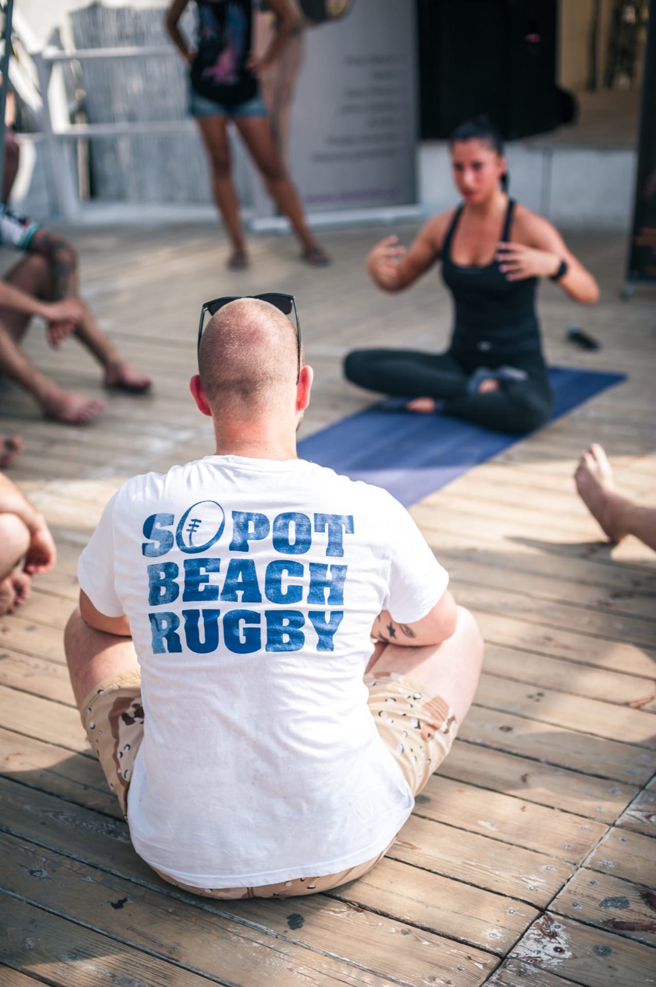 A group of people sit in a circle on a wooden terrace and participate in a yoga or meditation session. One person, with a shaved head and wearing a white shirt that reads "Sopot Beach Rugby," stands in the foreground, facing the woman leading the session. A photo essay from the fair captures this peaceful moment.  