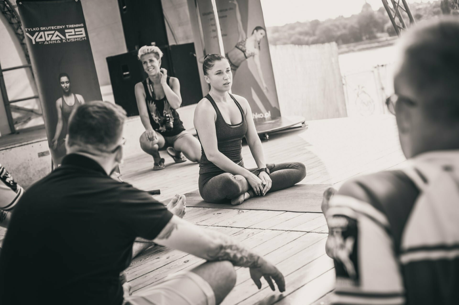 A group of people gathered on a wooden platform outside, immersed in a yoga or meditation session. A person in the middle sits with his legs crossed and leads the session. Other participants sit or kneel around and listen attentively. In the background you can see banners and a picturesque view, reminiscent of a photo fair.   