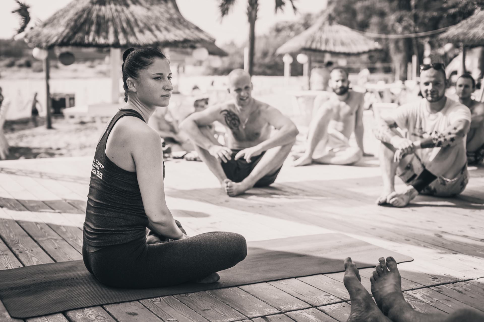 A woman sits with her legs crossed on a yoga mat and leads an outdoor yoga class. Around her on the mats sit several other people, some smiling. The setting is sunny, with thatched huts and palm trees in the background, suggesting a tropical or beach location - perfect for a photo fair.  