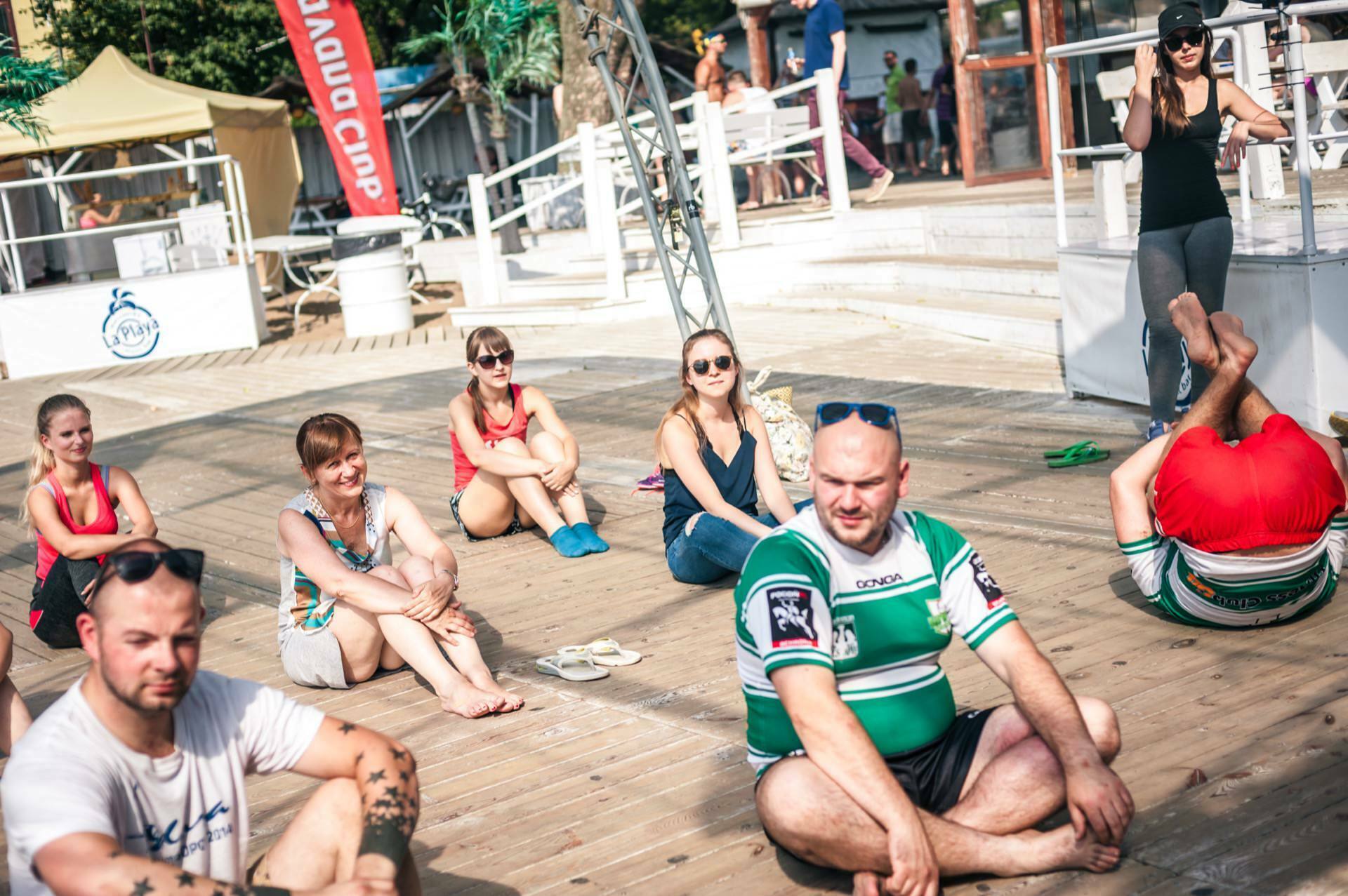 A group of people are sitting on a wooden terrace, apparently attending a yoga or fitness class. They are carelessly dressed, and one person is seen in a twisted yoga pose. A woman instructs at the front while others relax and listen. The scenery appears to be outdoors, perhaps captured as a photo essay of a trade show.   