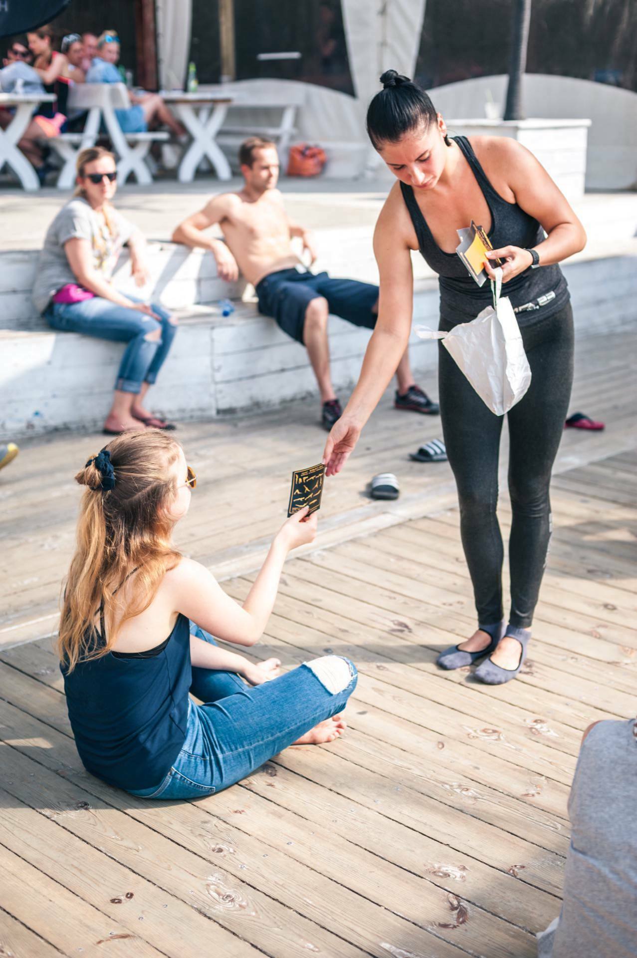 A woman in black attire hands a booklet to a seated blonde woman in a blue T-shirt and jeans on a wooden terrace, capturing the moment of a photo opportunity at the fair. Additional people sit on benches in the background and enjoy the sunny day. 