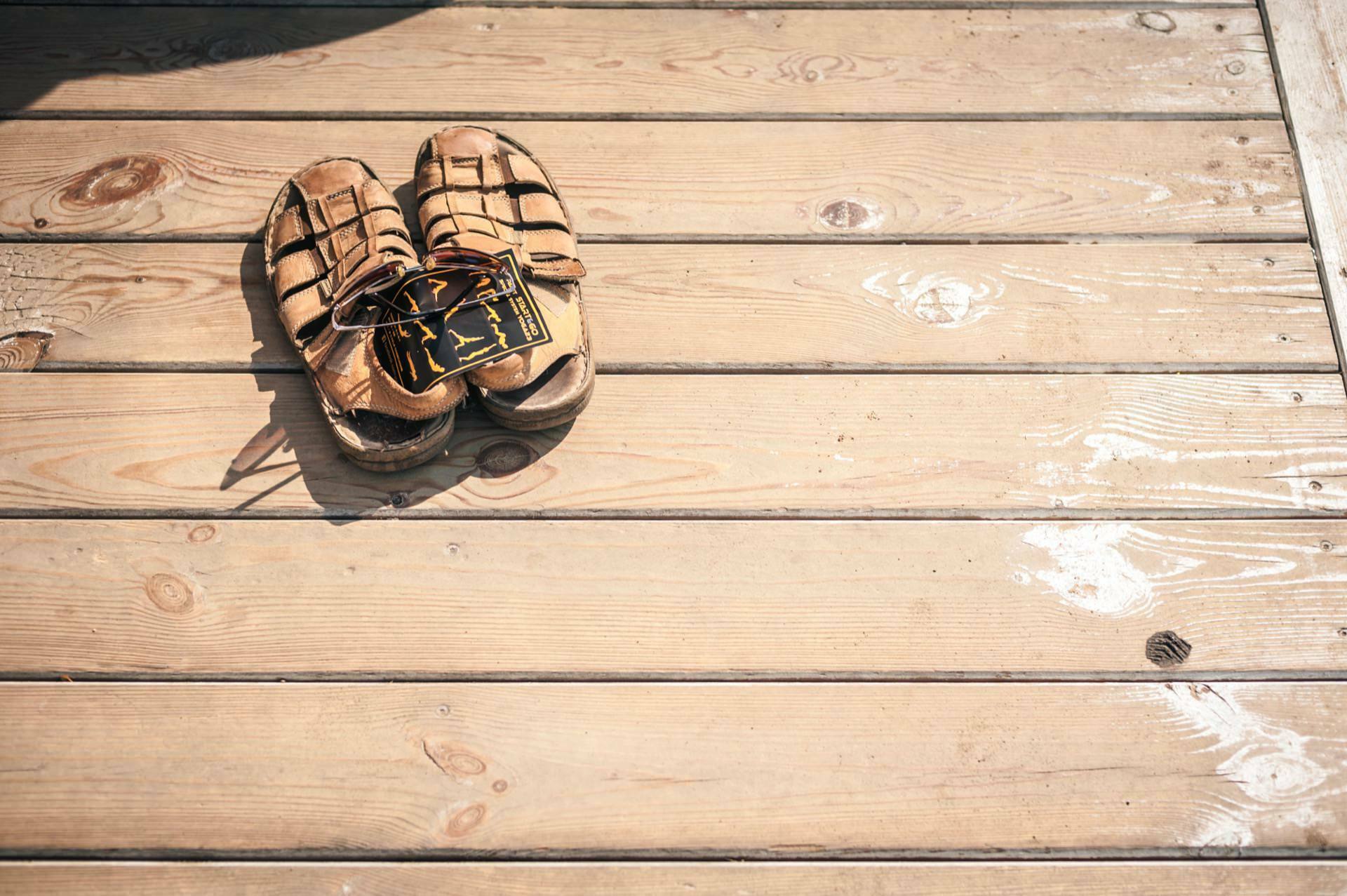 Worn leather sandals placed on a wooden deck with a pair of sunglasses. The deck has a faded, rustic look with visible knots and wood grains, evoking memories of a photo fair. The sunlit scene suggests a warm outdoor setting.  