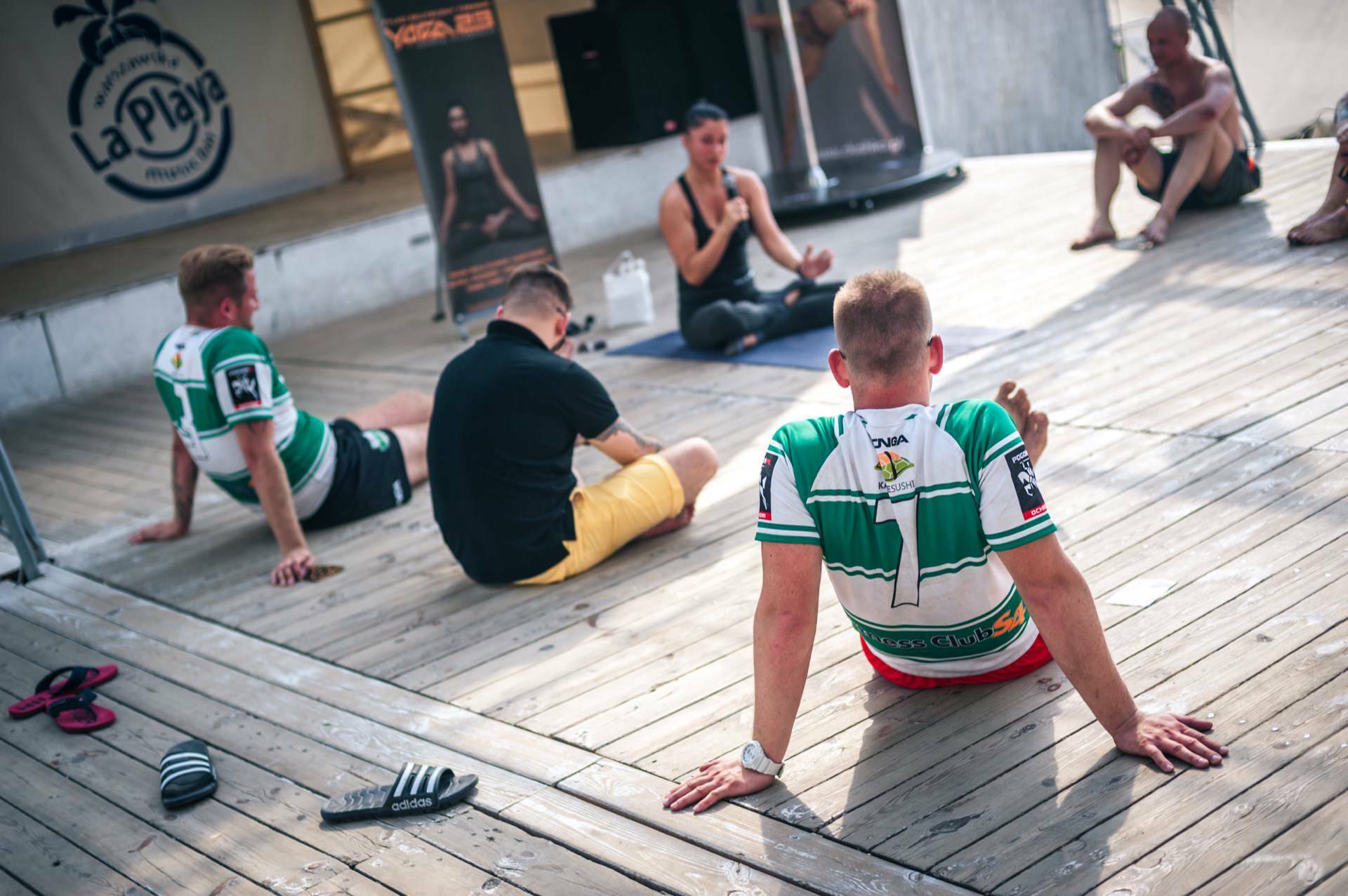 A small group of people sit on a wooden terrace and participate in an outdoor session led by an instructor. Two people in green and white sports jerseys sit in the foreground, while a black and white La Playa banner and palm tree decoration provide a vivid backdrop, creating a photo fair. 