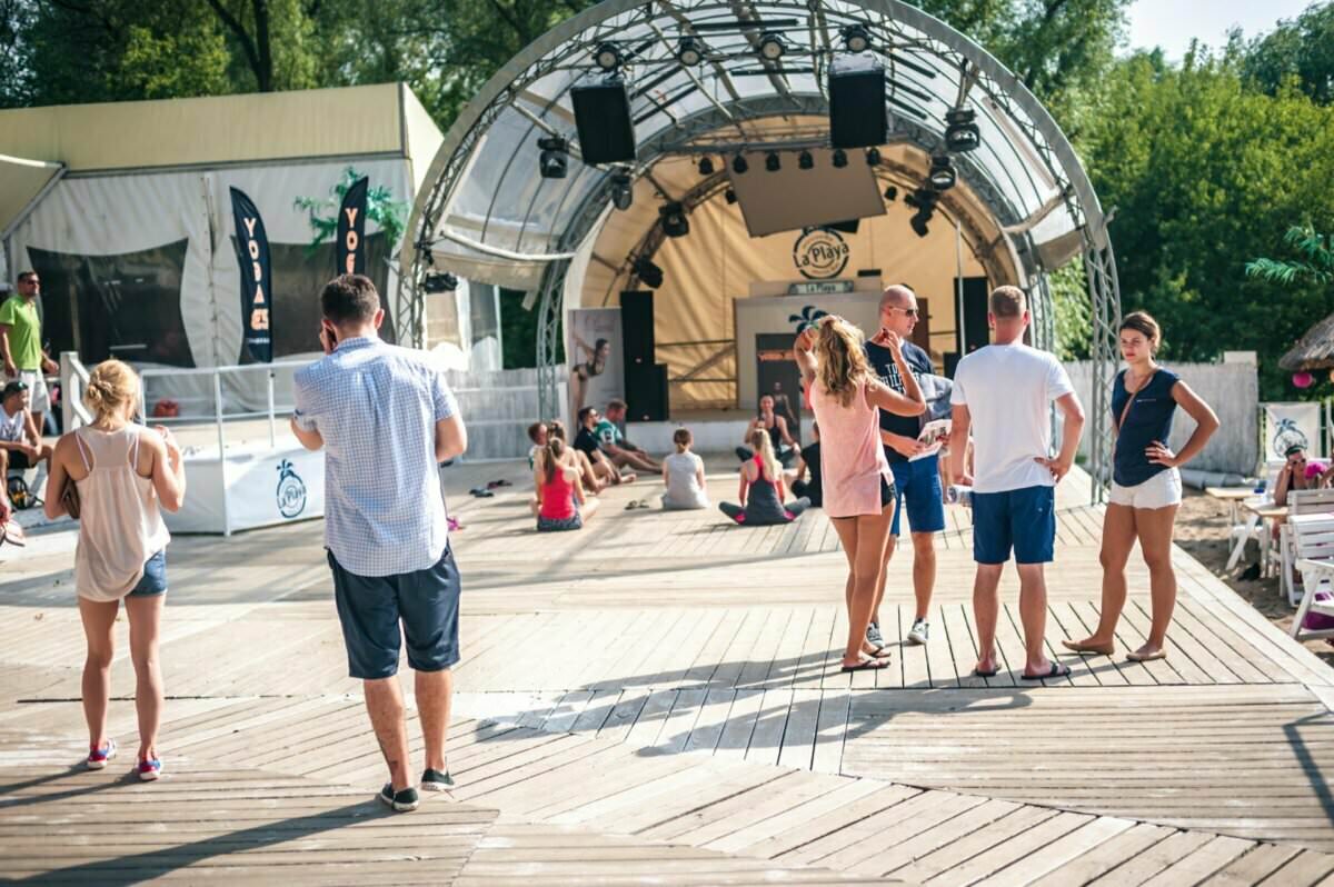 Grupa ludzi gromadzi się na drewnianym tarasie w miejscu imprezy plenerowej, tworząc ożywioną *fotorelację z targów*. Niektórzy stoją i rozmawiają, inni siedzą w tle. Scena z baldachimem jest wyraźnie widoczna, a drzewa zdobią odległy widok w ten słoneczny dzień.