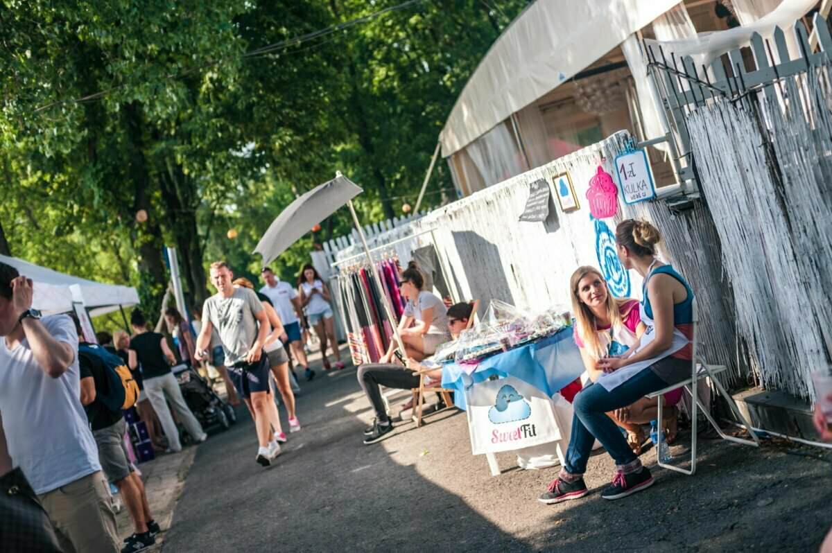 Tętniąca życiem scena targowa na świeżym powietrzu z ludźmi spacerującymi ścieżką. Dwie kobiety siedzą i rozmawiają przy stoisku z kolorowymi przedmiotami. Tabliczka na stole głosi „Słodkie Nadzienie”. Fotorelacja z targów uwiecznia inne stoiska i zieleń w tle pod czystym niebem, dodając tętniącej życiem atmosfery.