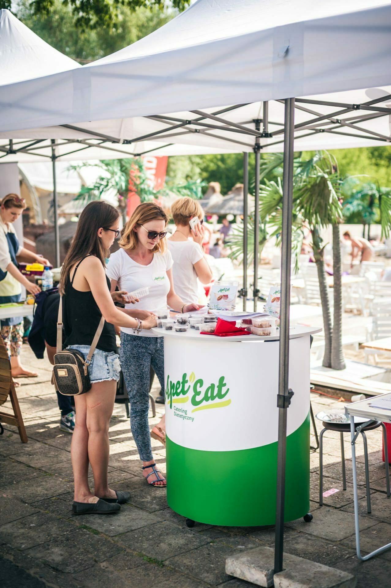 Two women stand at a booth with the "Sweet Eat" logo under a white tent at an outdoor event. One woman serves food samples, the other examines them. Other attendees and booths are visible in the background, in a sunny, green setting, capturing a vivid photo-op of the fair.  