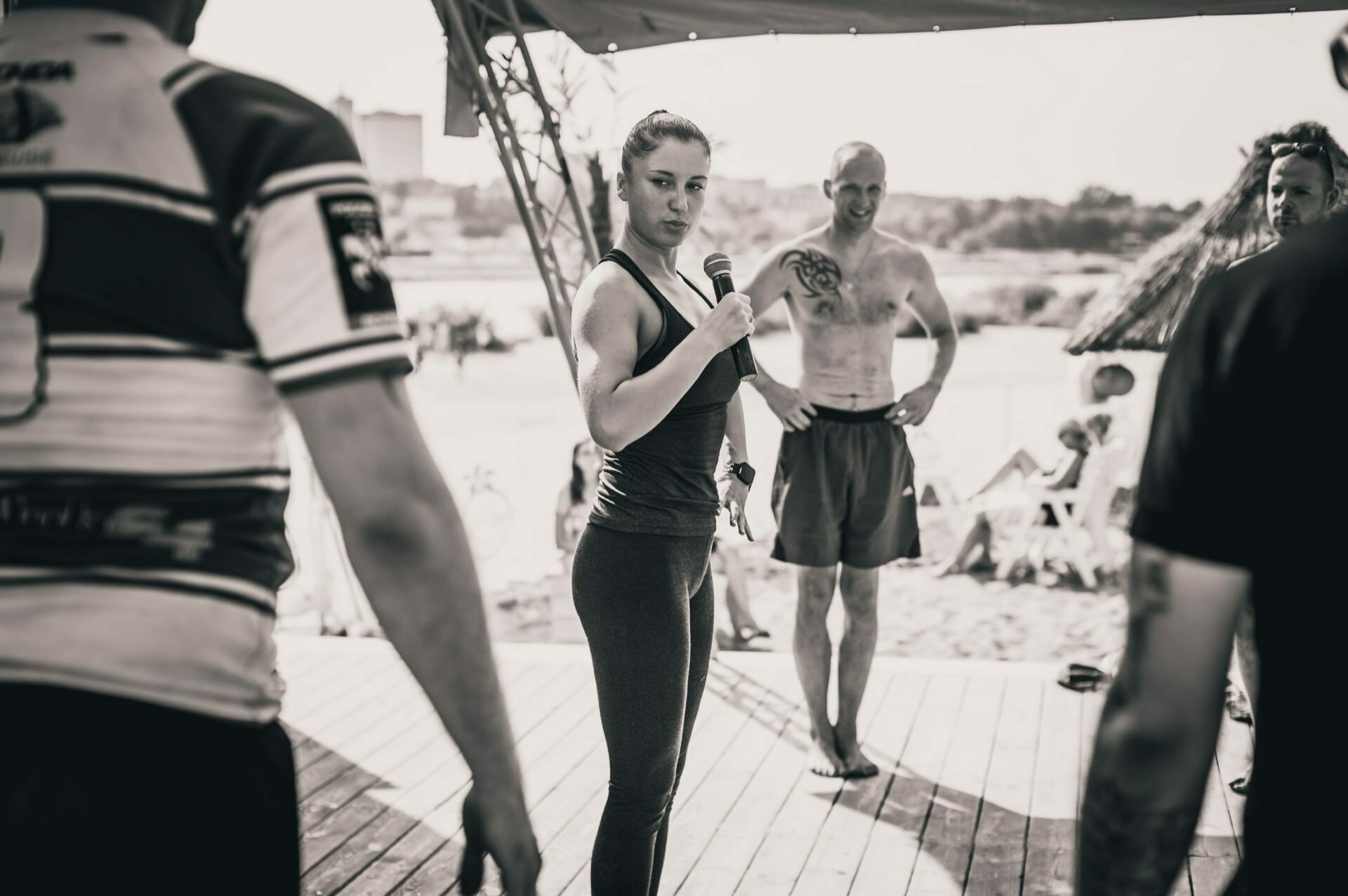 The woman holding the microphone stands on a stage by the beach and talks to the audience. She is dressed in sportswear, with her hair pulled back. Around her stand several men, one shirtless and with a tattoo. Beachgoers relax in the background under thatched umbrellas. The photo report from the fair perfectly captures the vibrant atmosphere.    