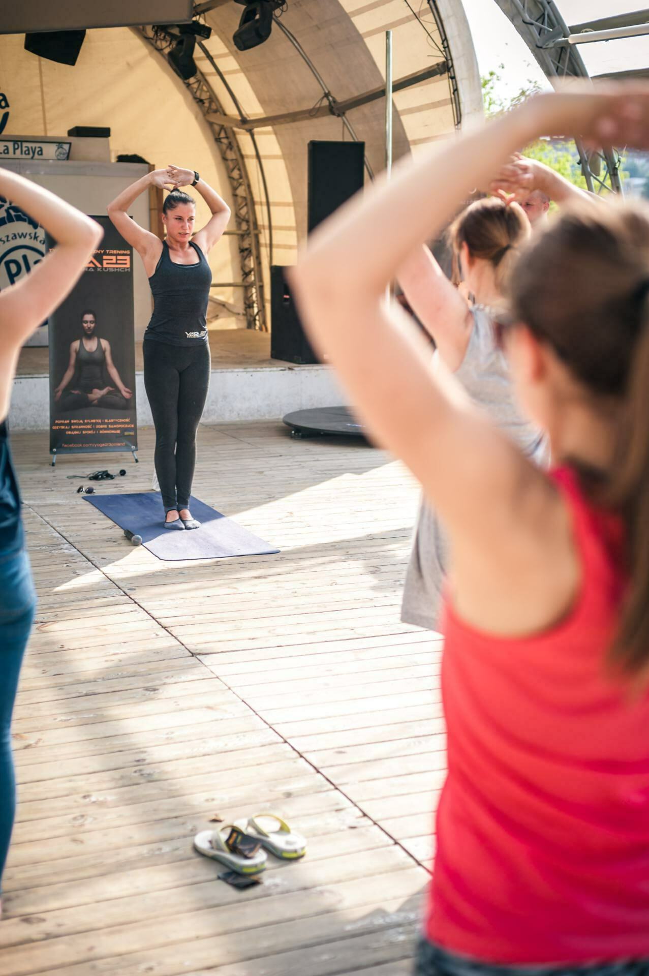 A group of people participate in an outdoor yoga class on a wooden terrace. The instructor, standing on a yoga mat, demonstrates a pose with arms raised and hands entwined behind the head. Participants follow along, imitating the pose. Shoes are placed nearby. It's like a quiet photo essay of a wellness activity fair.    