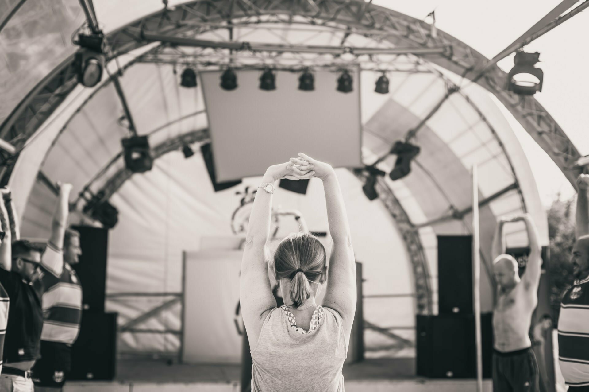 A group of people outdoors participate in a yoga or stretching class under a large dome-shaped structure. The scene is in black and white and resembles a photo fair, with the instructor leading from the stage while participants raise their arms and stretch. 