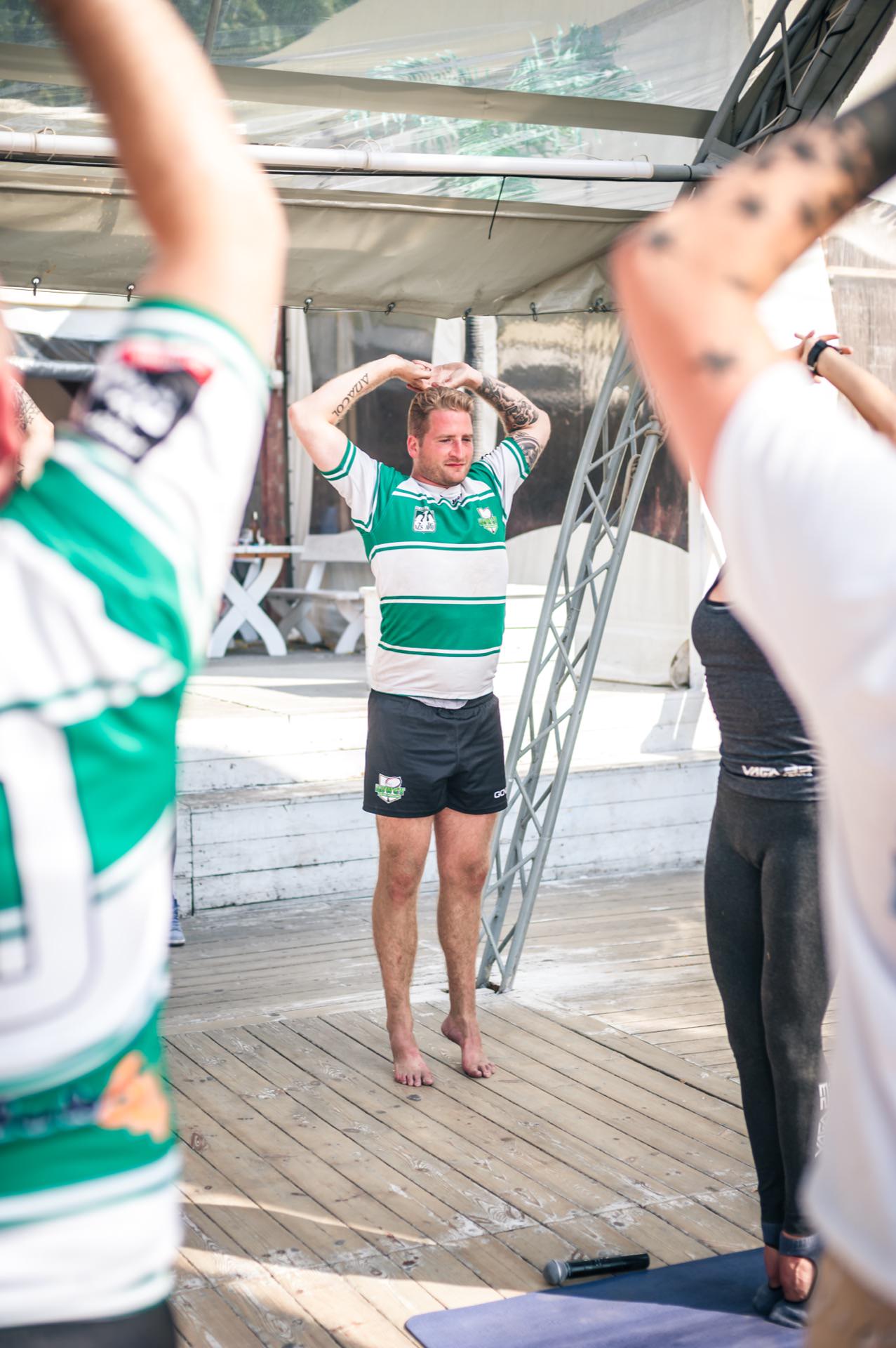 A man in a green and white sports shirt and black shorts stands barefoot on a wooden terrace, with his arms crossed and hands resting on his head. Surrounded by other people in exercise positions, it is clear that the Photo Fair captures an engaging group activity, likely taking place outdoors under a canopy. 