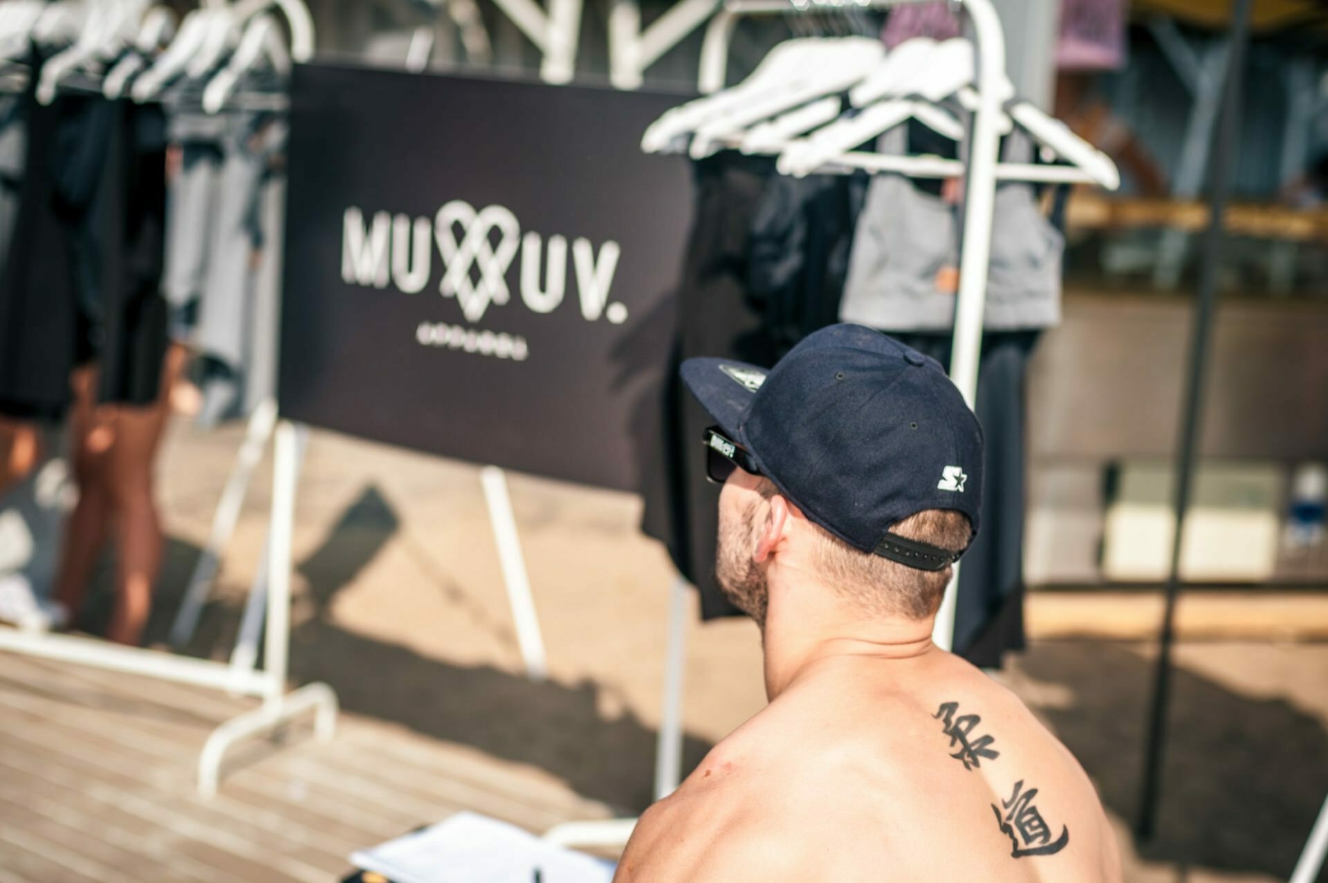 A shirtless man with tattoos, wearing a black cap and sunglasses looks at a clothing display with the caption "MUUV CLOTHING." A variety of clothes are hung on hangers in the display, and the setting seems to be an outdoor market or a photo-op. 