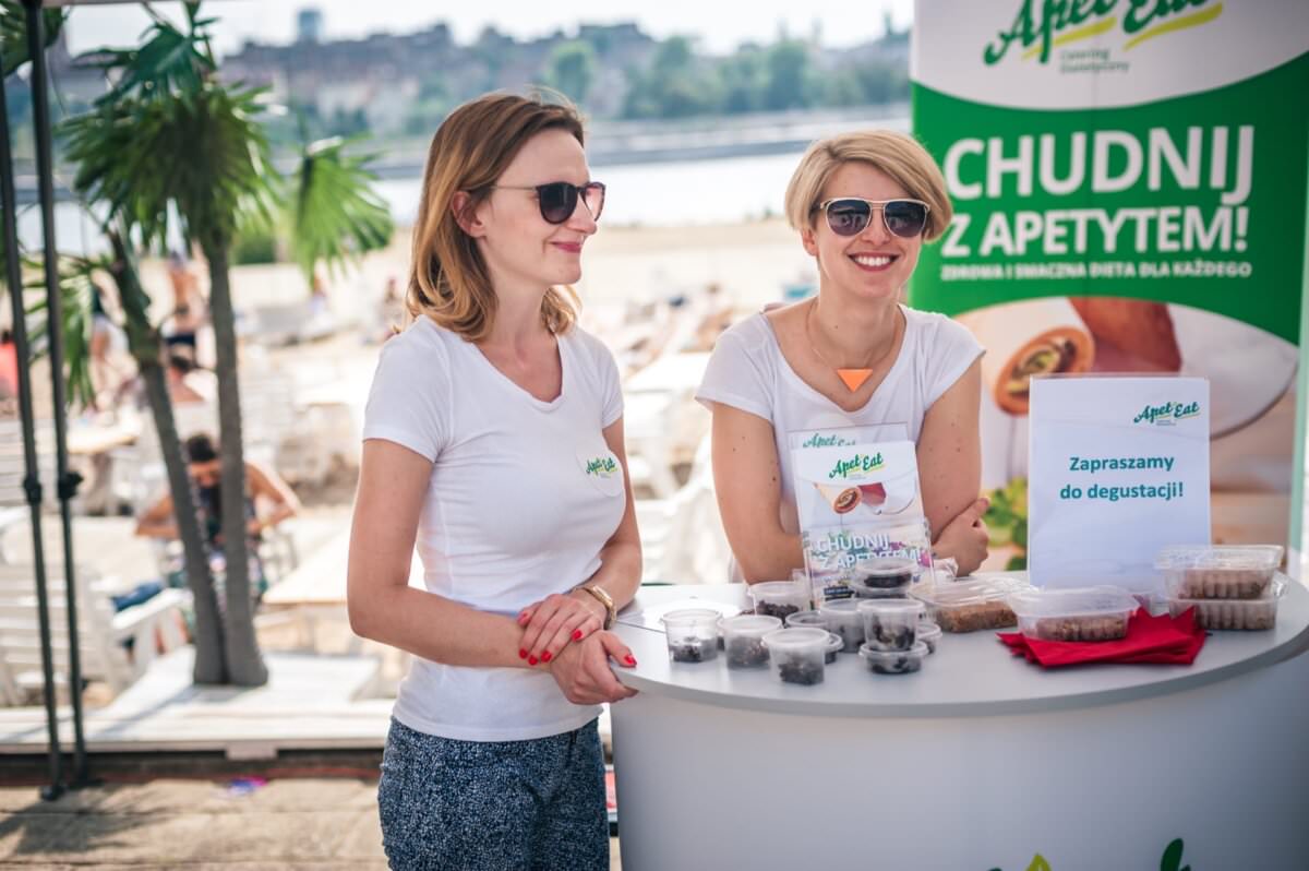 Dwie uśmiechnięte kobiety stoją na stoisku promocyjnym przy plaży i kupują produkt zdrowotny. Noszą białe koszulki z zielonym logo i okulary przeciwsłoneczne. Na stole znajdują się różne próbki w małych filiżankach oraz tabliczka zachęcająca do degustacji. Tło przedstawia słoneczną plażę, idealną do tej fotorelacji z targów.