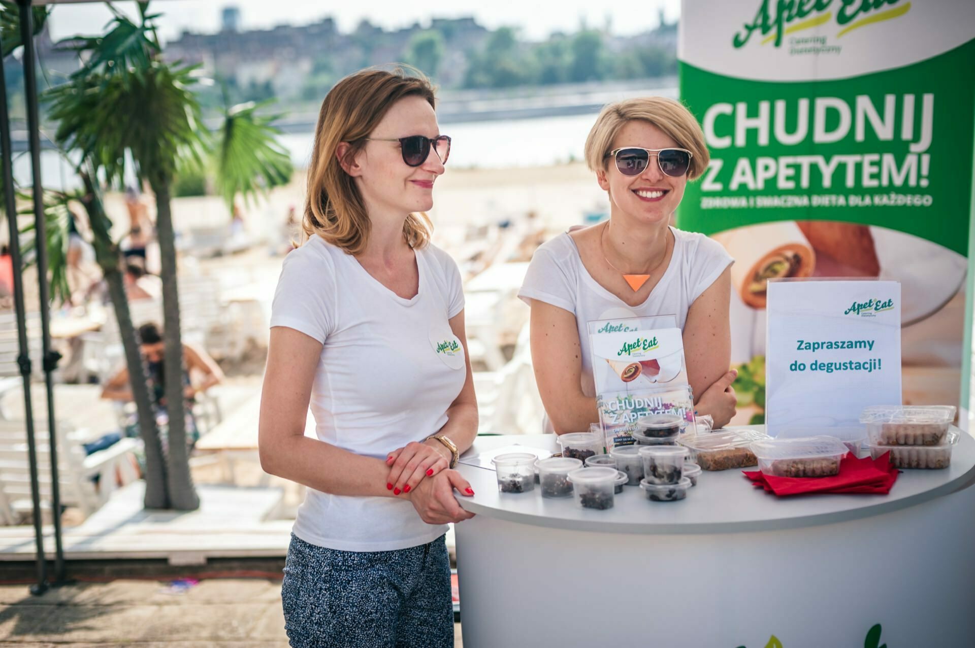 Two smiling women stand at a promotional booth by the beach and buy a health product. They wear white T-shirts with green logos and sunglasses. On the table are various samples in small cups and a sign inviting them to taste. The background shows a sunny beach, perfect for this photo fair.   