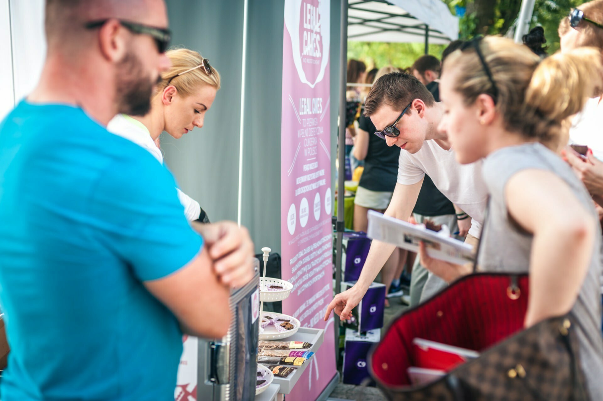 People at an outdoor stand are checking out and buying items. A man in a white shirt points to products on a table, and a vendor explains. Another man in a blue shirt stands nearby, and a woman holds a red bag. The atmosphere is lively and busy - a true photo-op of the fair.   