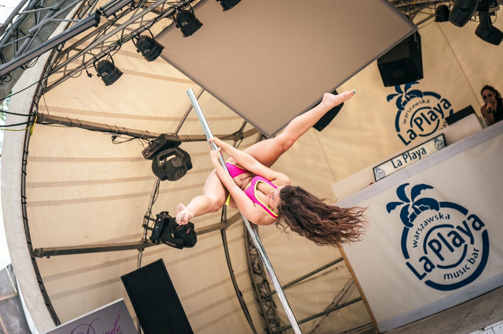 A woman in a bright pink outfit performs twine in the air on a pole during an outdoor event under a large tent. The stage is equipped with lighting and speakers, and a banner with "La Playa" branding is visible in the background. One more person appears at the DJ booth in this exciting Photo Report from the fair.  