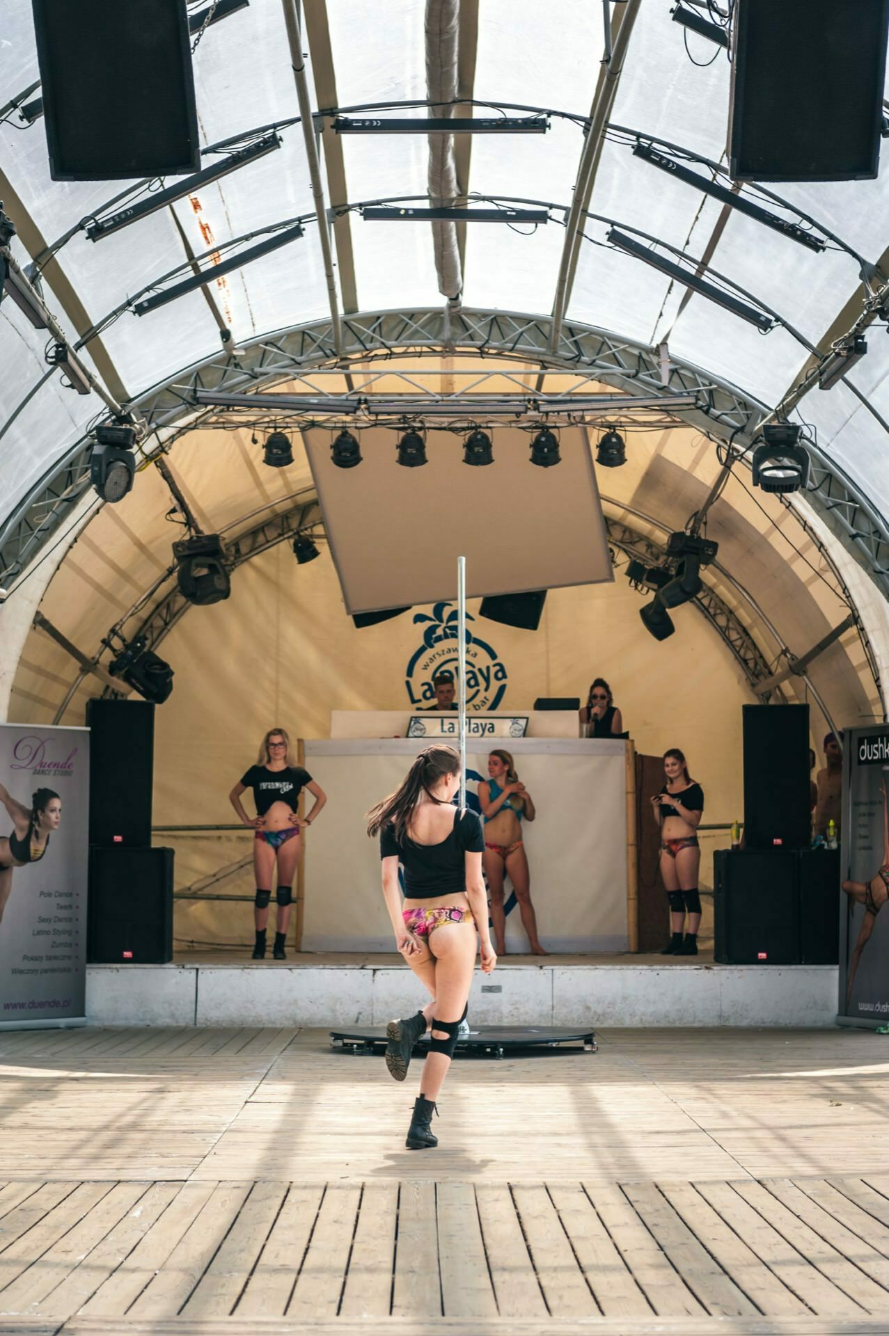 A woman performs a pole dance on a stage with a curved tent-like roof. She is surrounded by several other women standing in the background, as well as speakers and banners. In a vibrant photo essay from the fair, additional lighting and sound equipment can be seen suspended above the stage.  