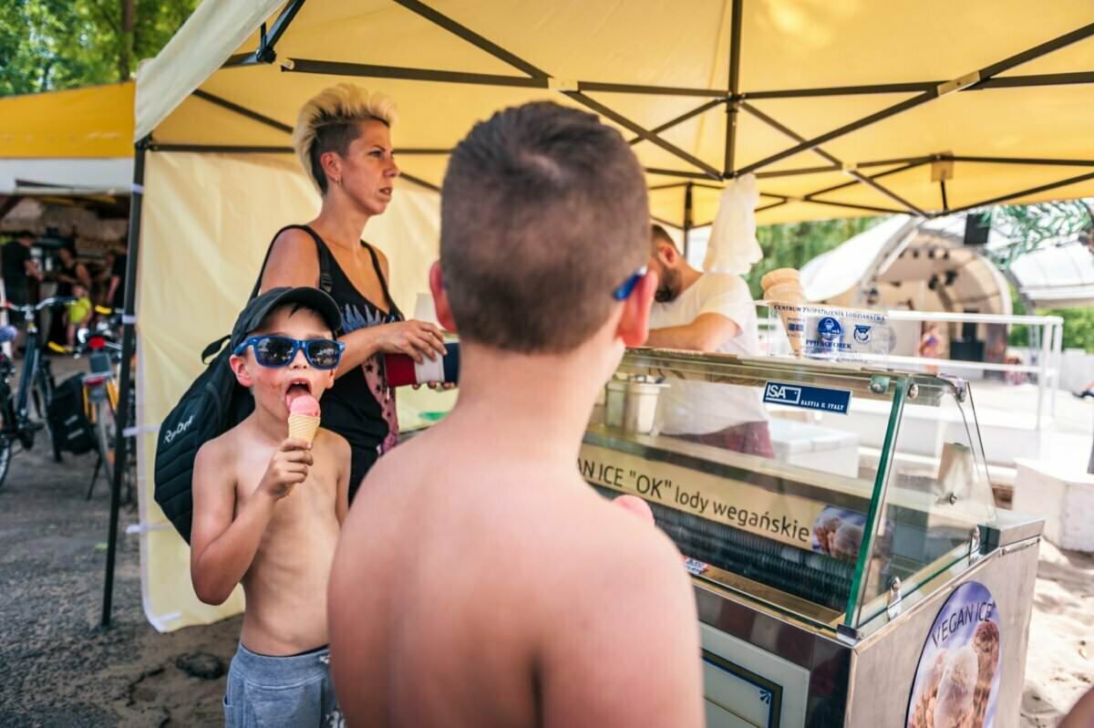 Młody chłopiec w okularach przeciwsłonecznych zajada się lodami, stojąc bez koszulki obok wózka z lodami pod żółtym namiotem. Inny chłopiec bez koszulki stoi na pierwszym planie, a za nimi stoi kobieta z krótkimi włosami. W tle widać inne osoby, które oddają klimat Fotorelacji z targów.